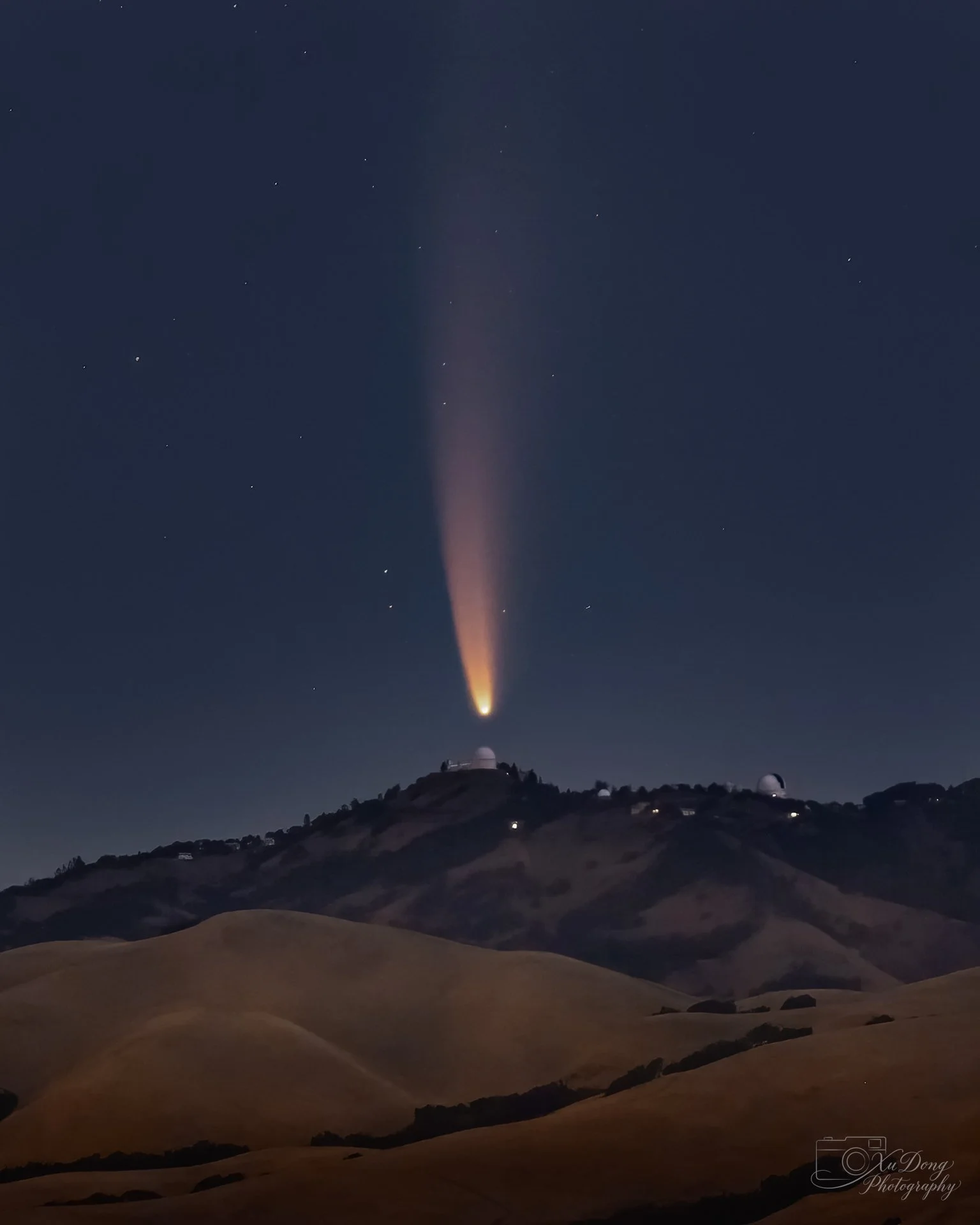 A rare celestial capture of Comet NEOWISE displaying its brilliant tail across the night sky, positioned above a mountain-top observatory at dawn.