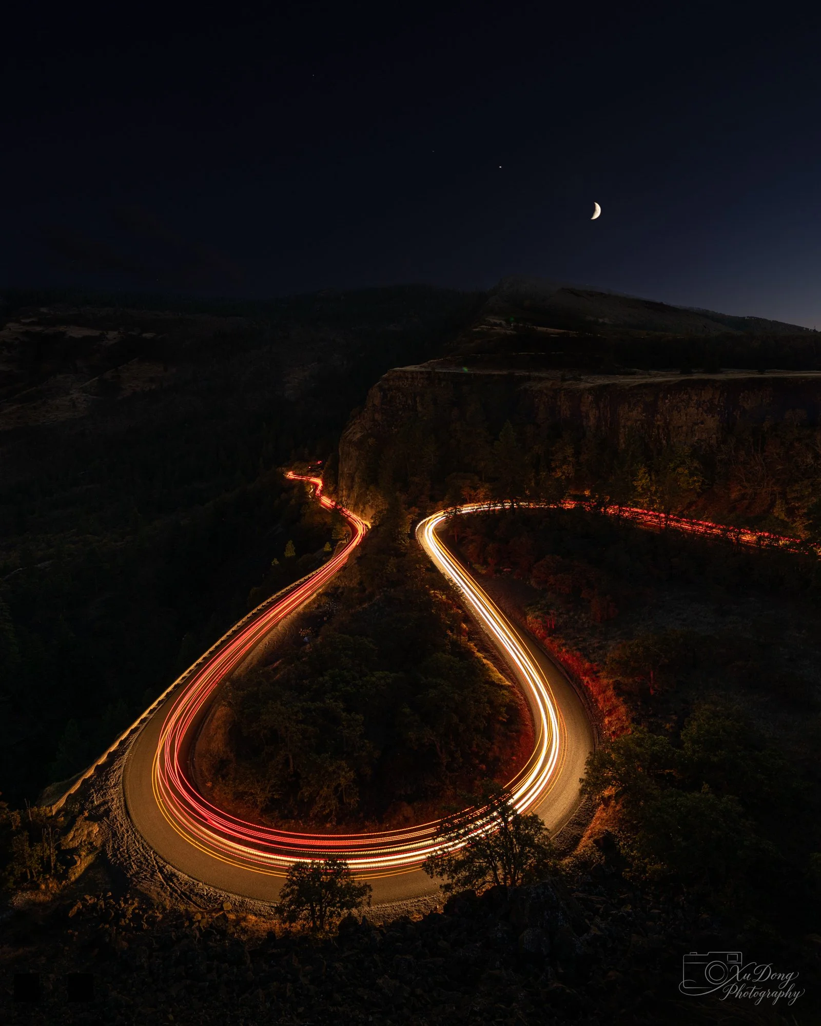 A dynamic night photography composition featuring a winding mountain road with long-exposure vehicle light trails that mirror the curve of the moon above.