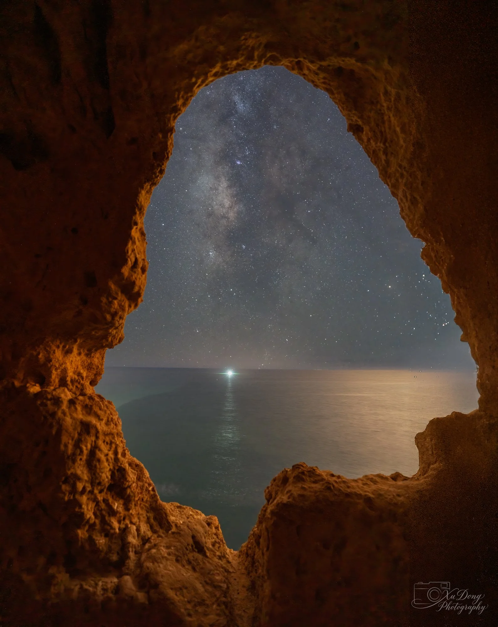 A unique perspective from within a rugged limestone sea cave in Southern Portugal, framing the vast, star-filled night sky and the glowing core of the Milky Way over the Mediterranean Sea.