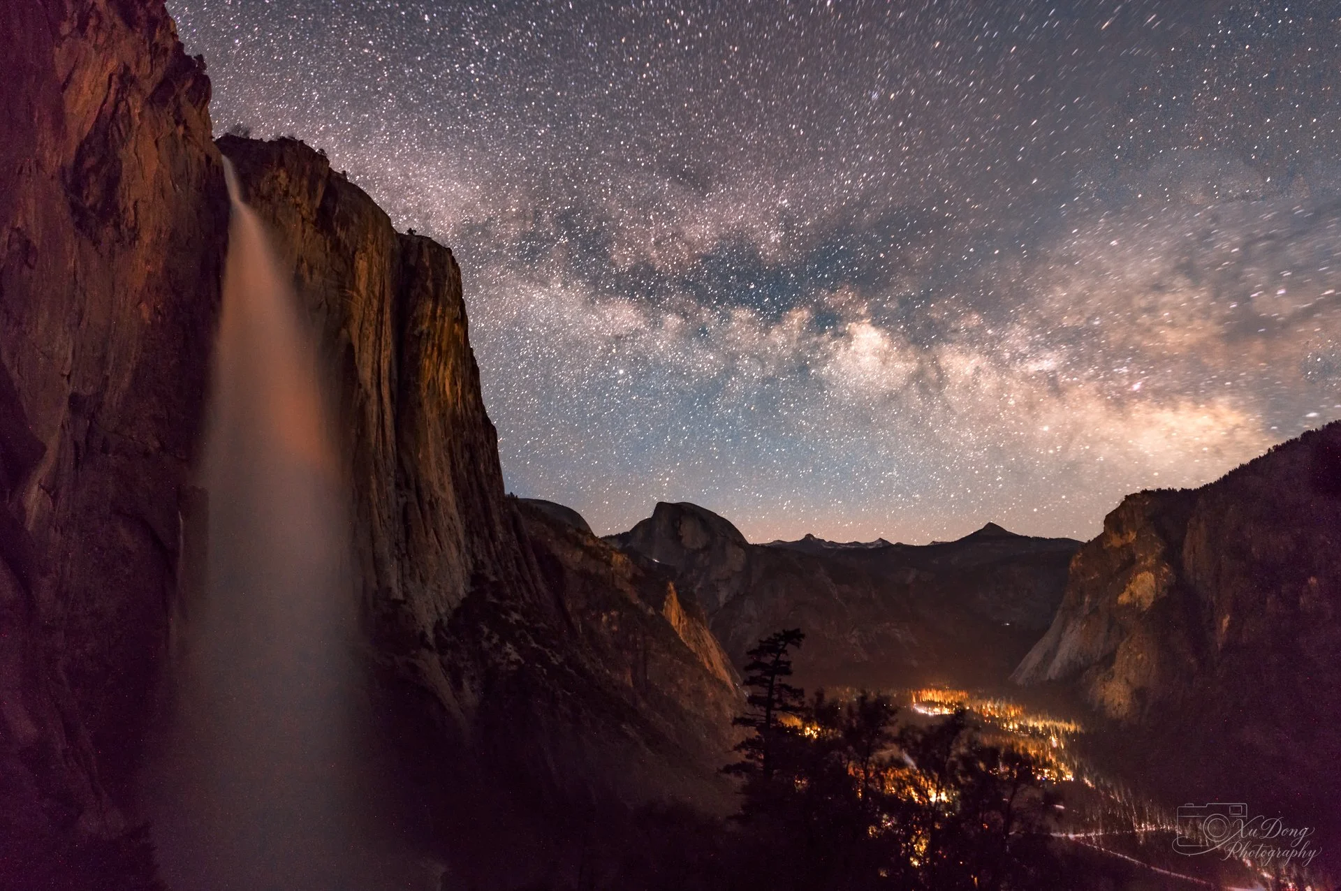 A majestic nightscape of the Yosemite Valley, featuring the Milky Way galaxy arching over the towering granite cliffs and the illuminated silhouette of Upper Yosemite Fall
