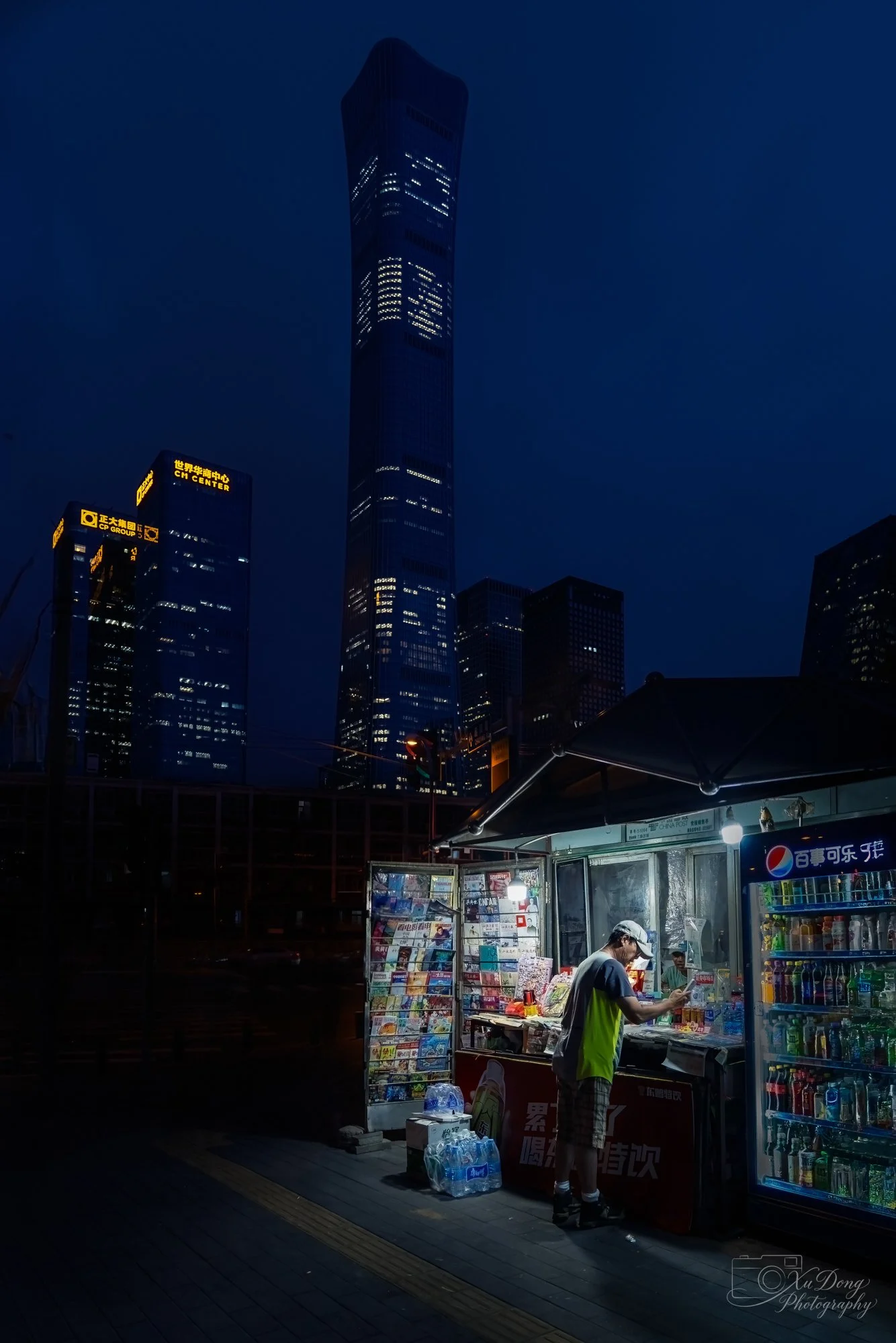 The human scale of a Beijing street-side newsstand glowing in the evening, contrasted by the towering, cold blue skyscrapers of the CBD.