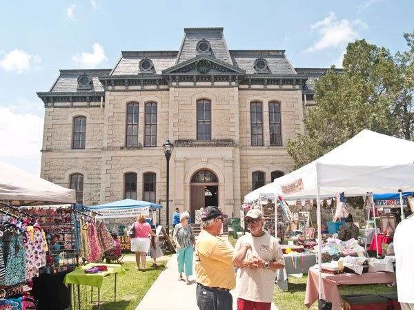 People browsing outdoor market stalls in front of the historic Blanco Courthouse with arched windows and a central door.