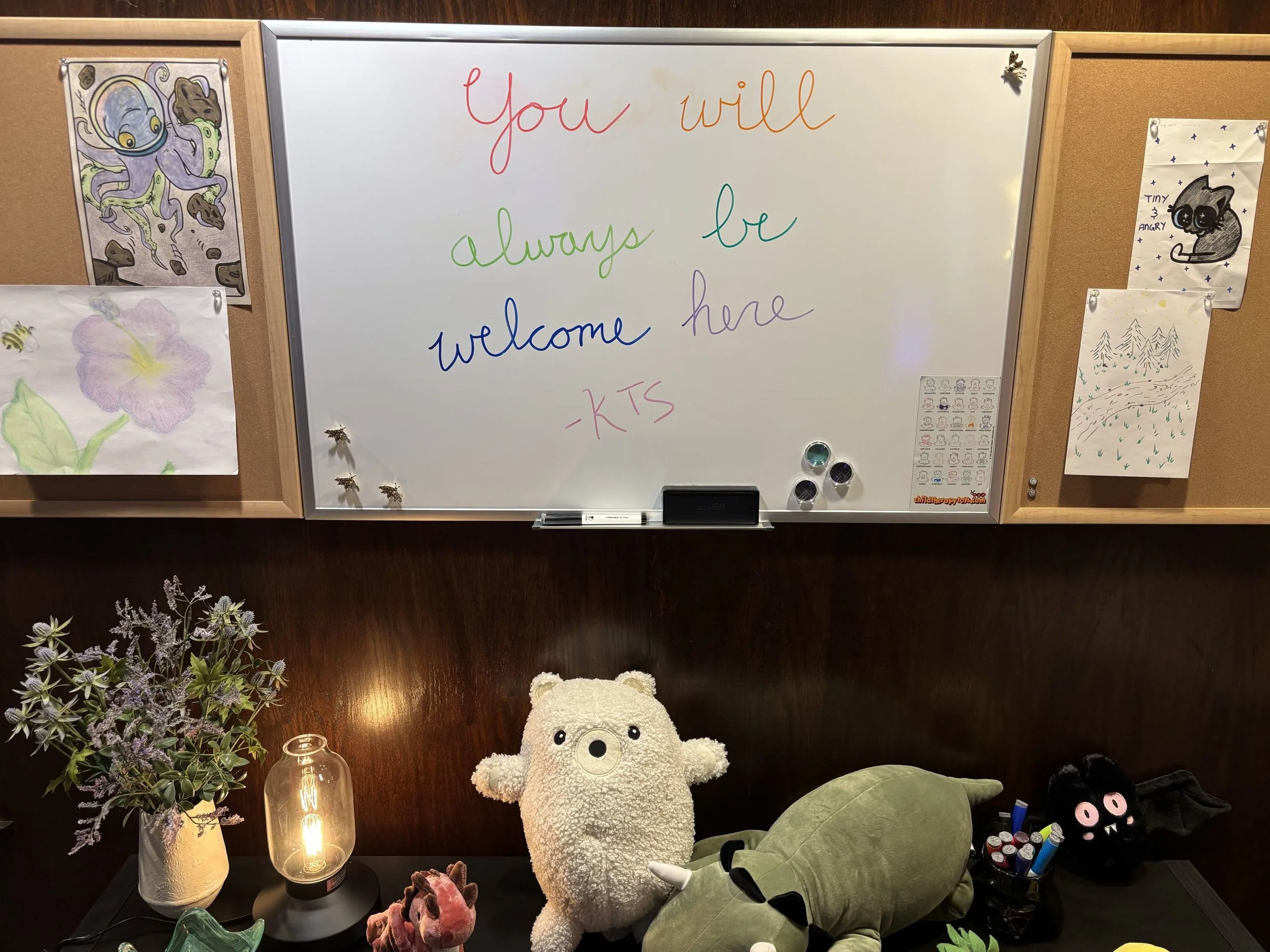 A photo of a therapy office's whiteboard reading "You Will Always Be Welcome Here - KTS" in rainbow colors