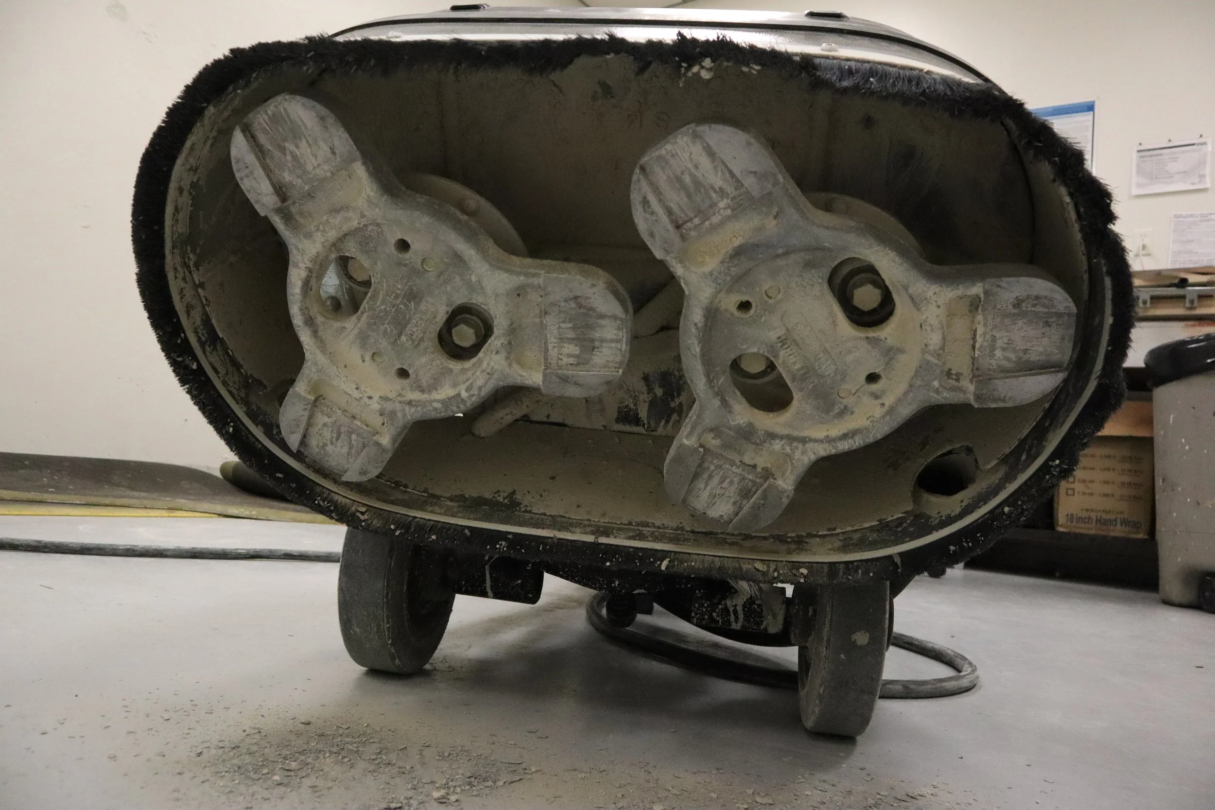 The underside of a robotic lawn mower showing its cutting blades and wheels, with a dusty floor and some workshop tools and items in the background.