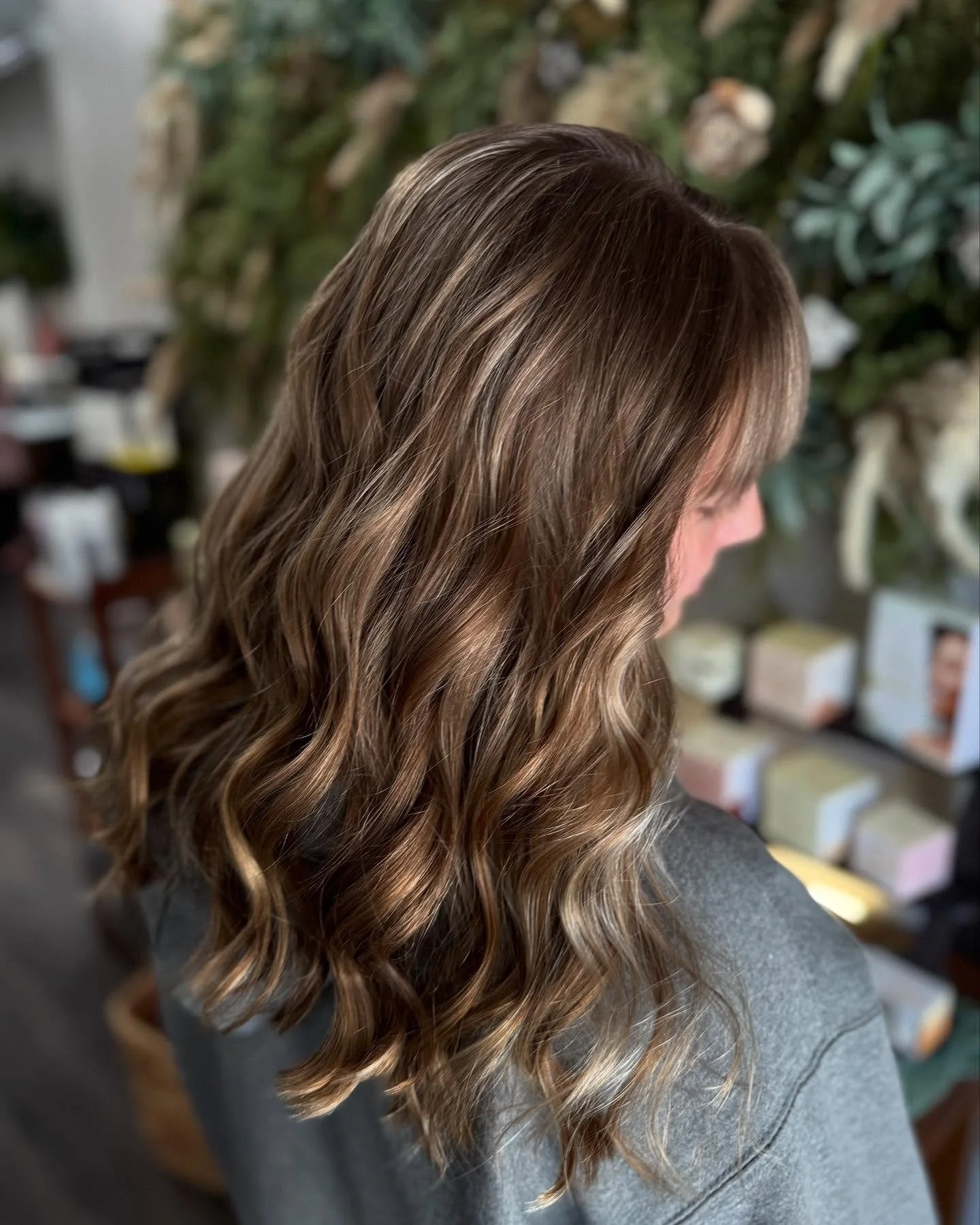 Side view of a woman with shoulder-length wavy light brown hair with highlights, wearing a gray top, in a salon or similar setting.