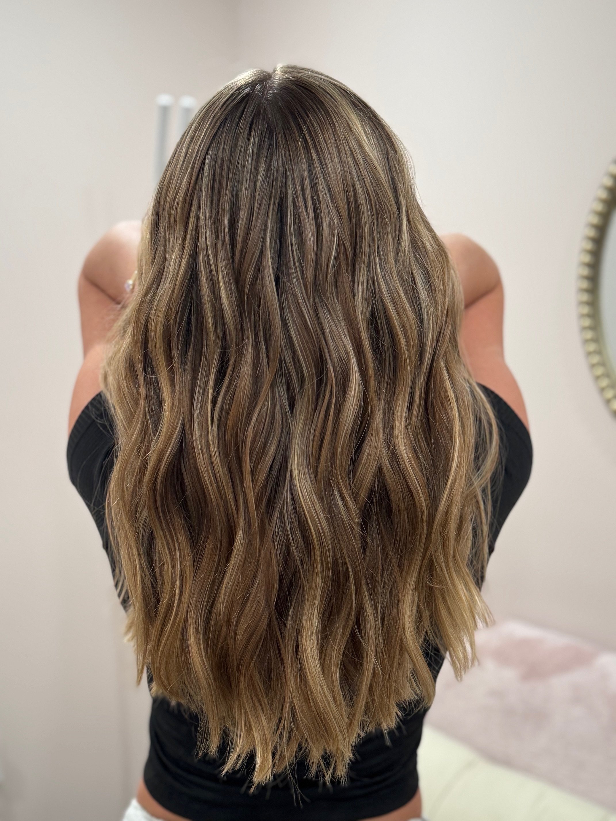 Back view of a woman with long, wavy, light brown hair standing in a room with a mirror on the wall.