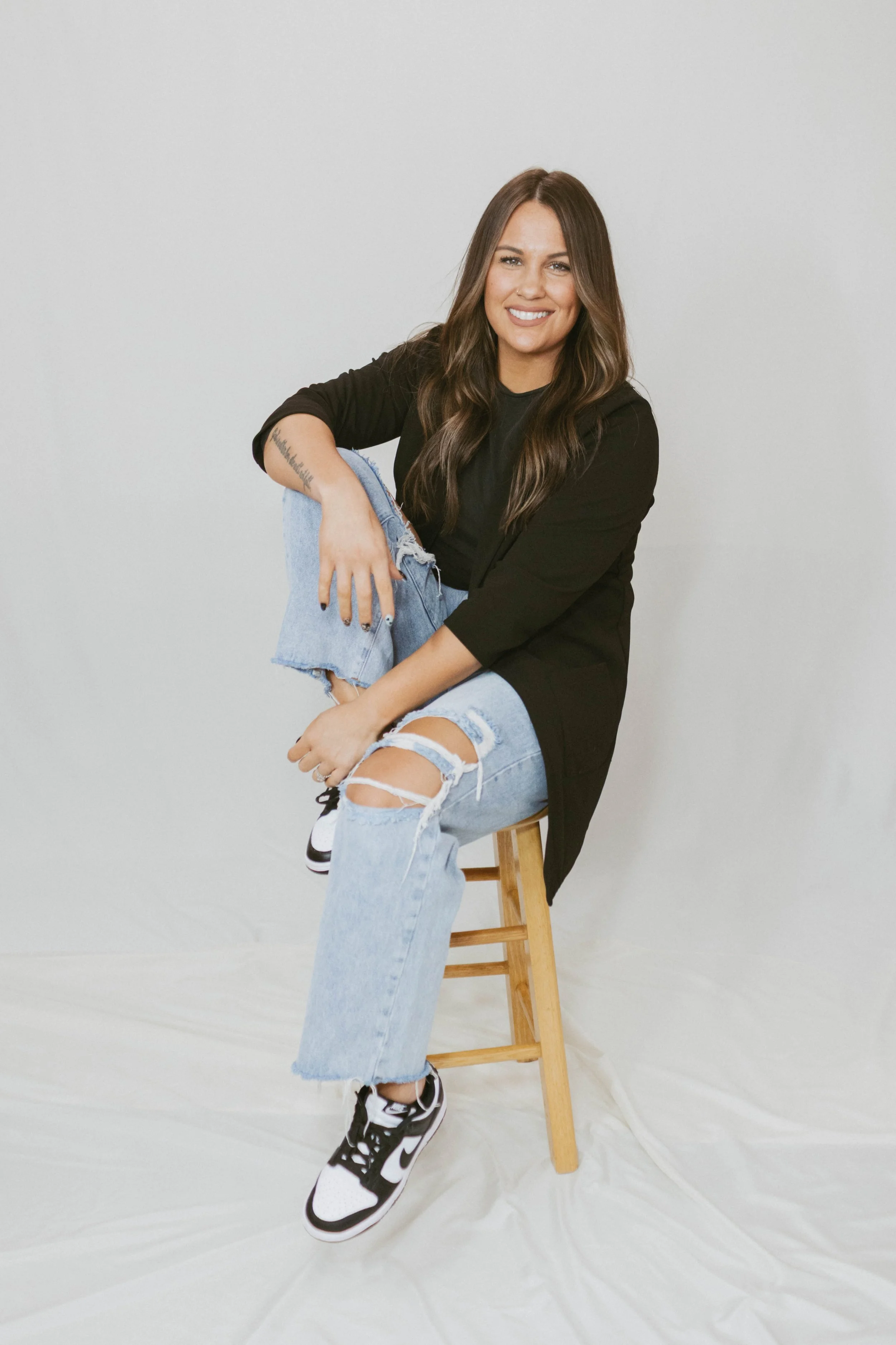 A young woman with long brown hair, smiling, sitting on a wooden stool against a plain white background. She is wearing a black long sleeve shirt, ripped light blue jeans, and black and white sneakers.