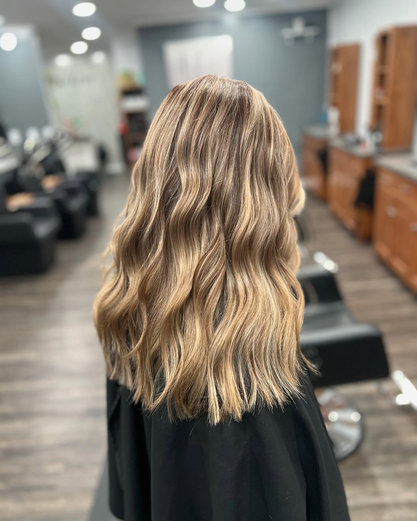 Back view of a woman with long, wavy, blonde hair in a salon setting.