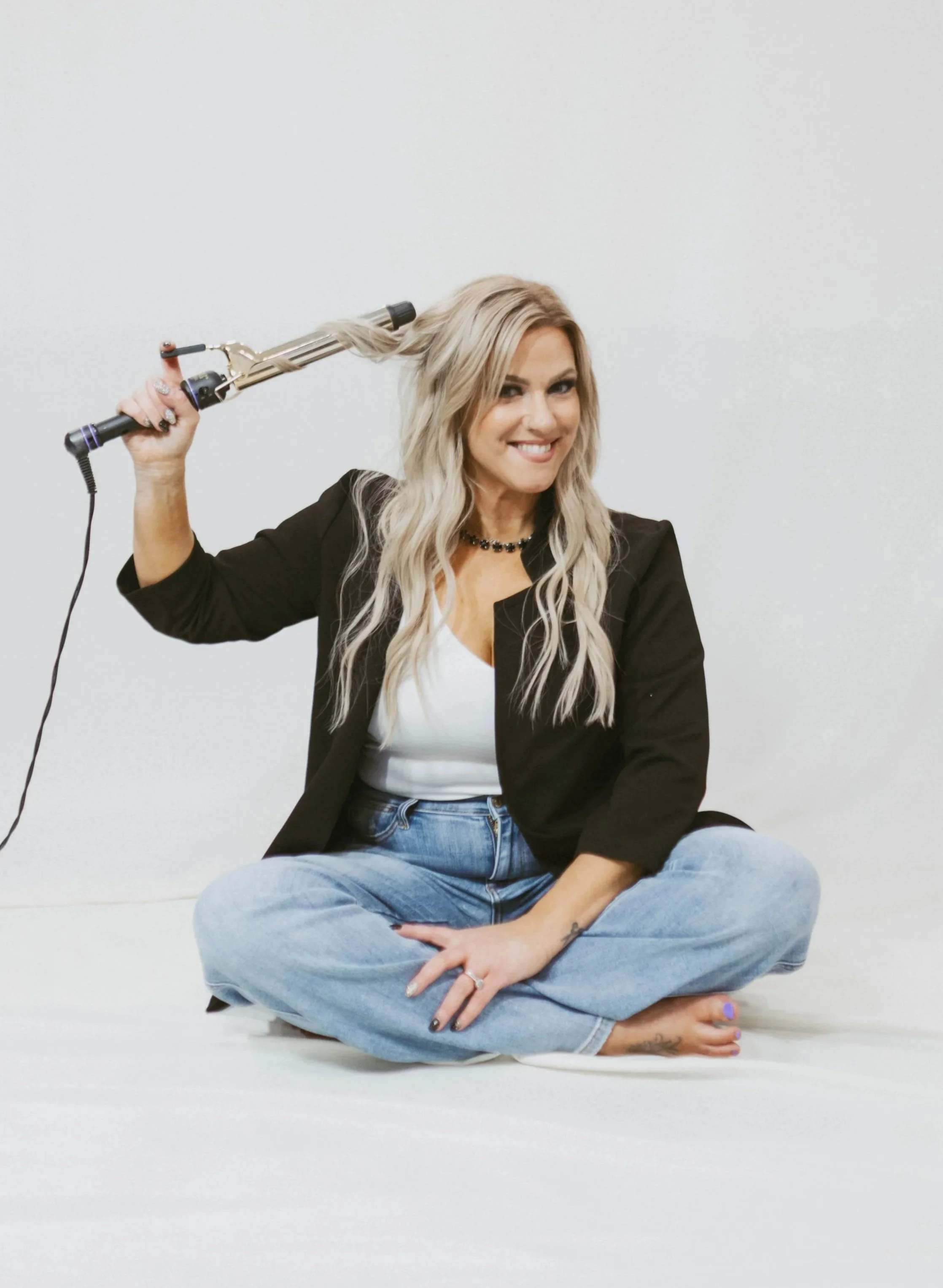 Woman sitting cross-legged on the floor, smiling, holding a curling iron in her right hand, against a plain white background.