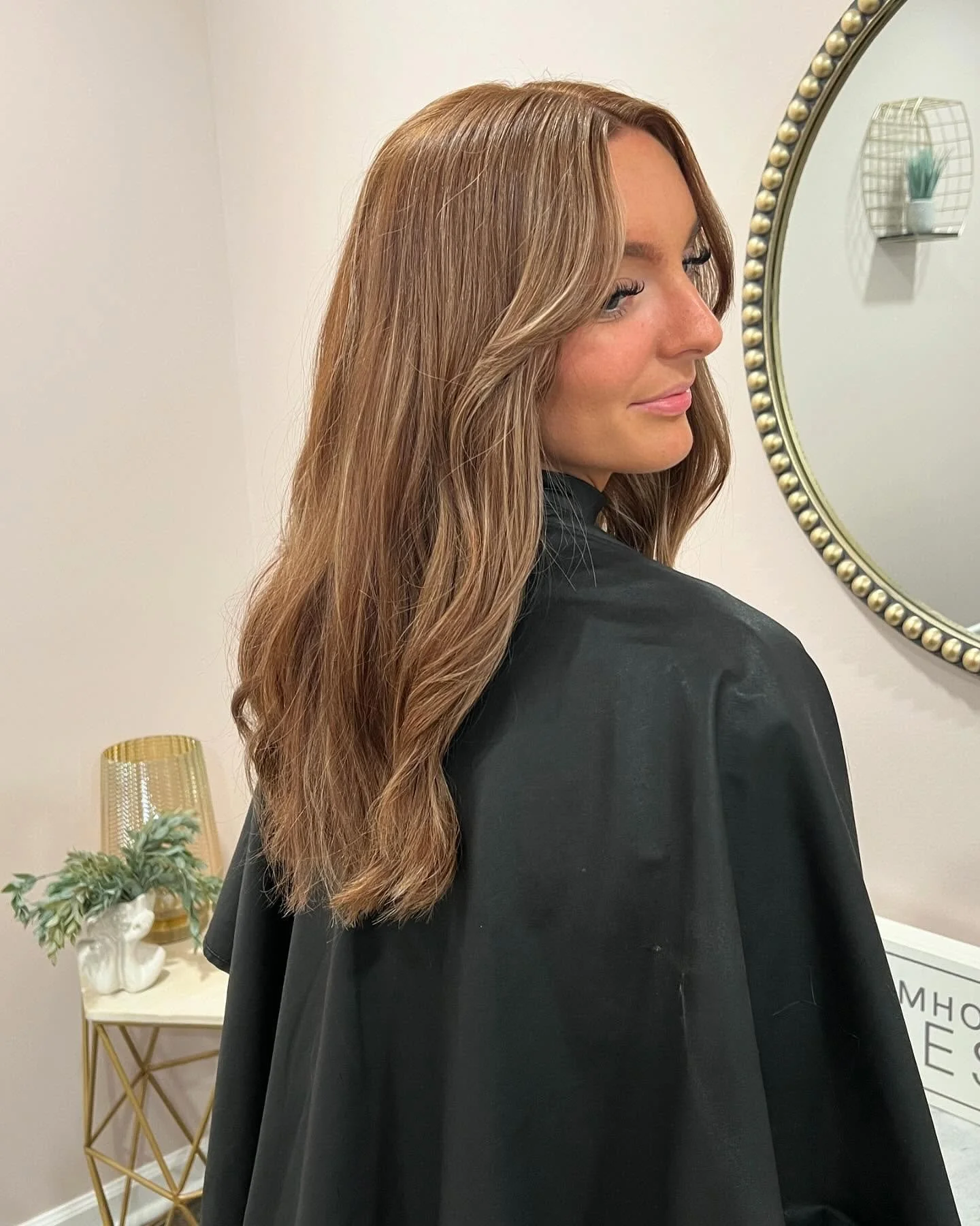 A woman with light brown, wavy hair looking to the side in front of a mirror, wearing a black cape or coat.