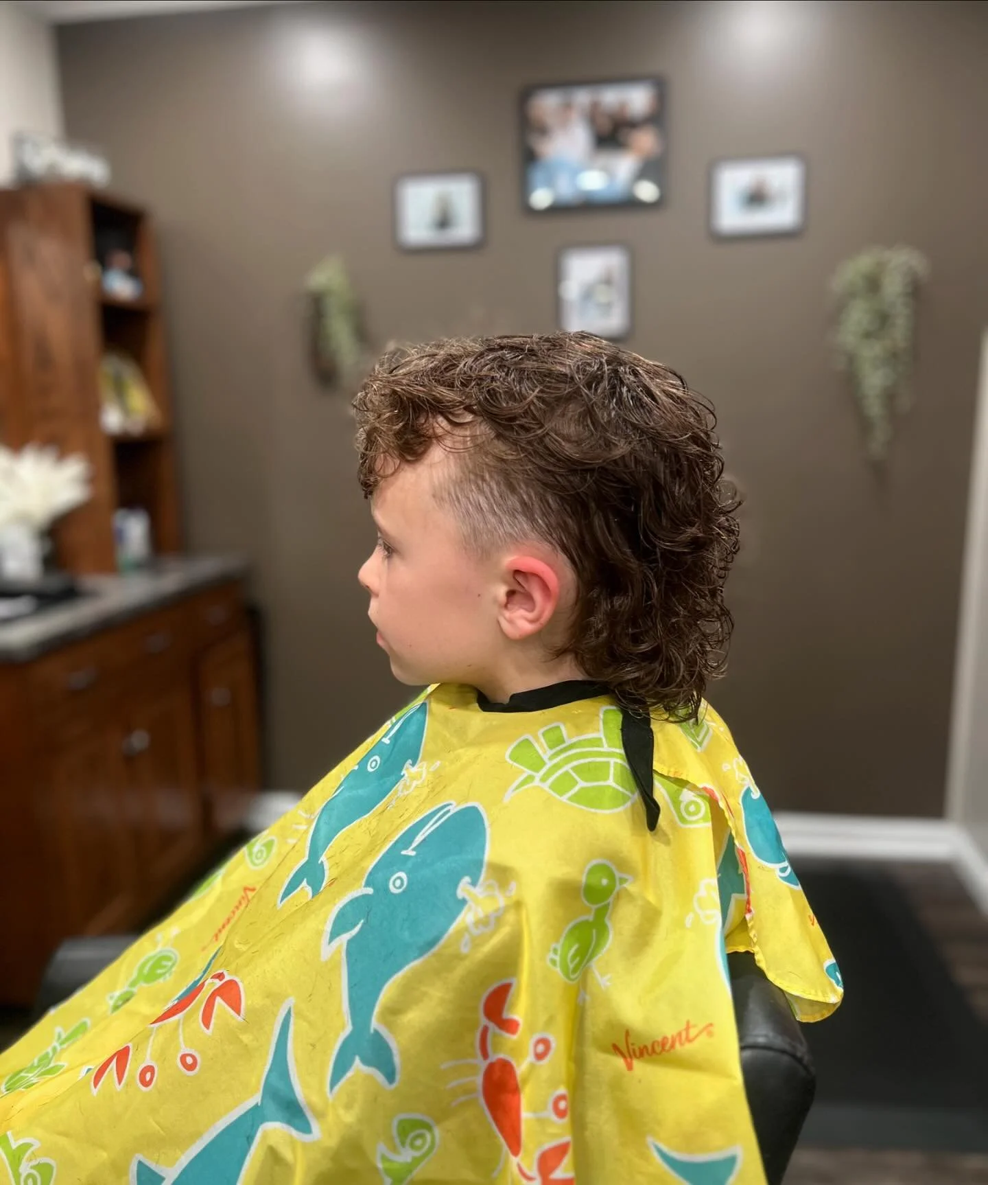 Young boy with a curly mullet hairstyle sitting in a salon chair, wearing a colorful cape with fish patterns, in a salon with framed pictures and wall decorations.