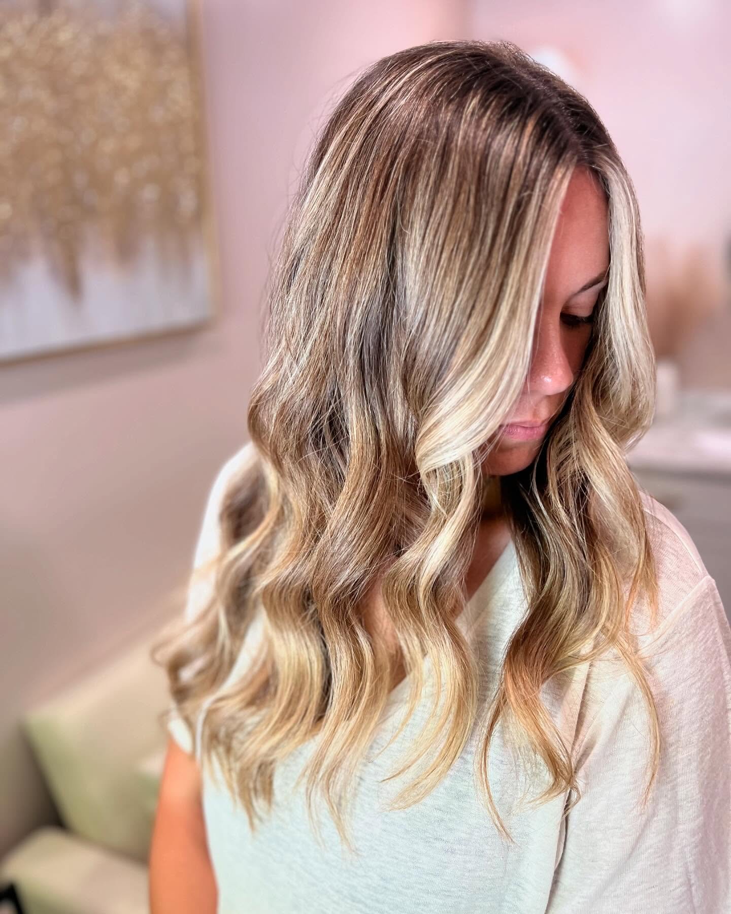 A woman with long, wavy blonde hair with highlights, looking down, sitting on a bed with a pink background and a gold wall art in the background.