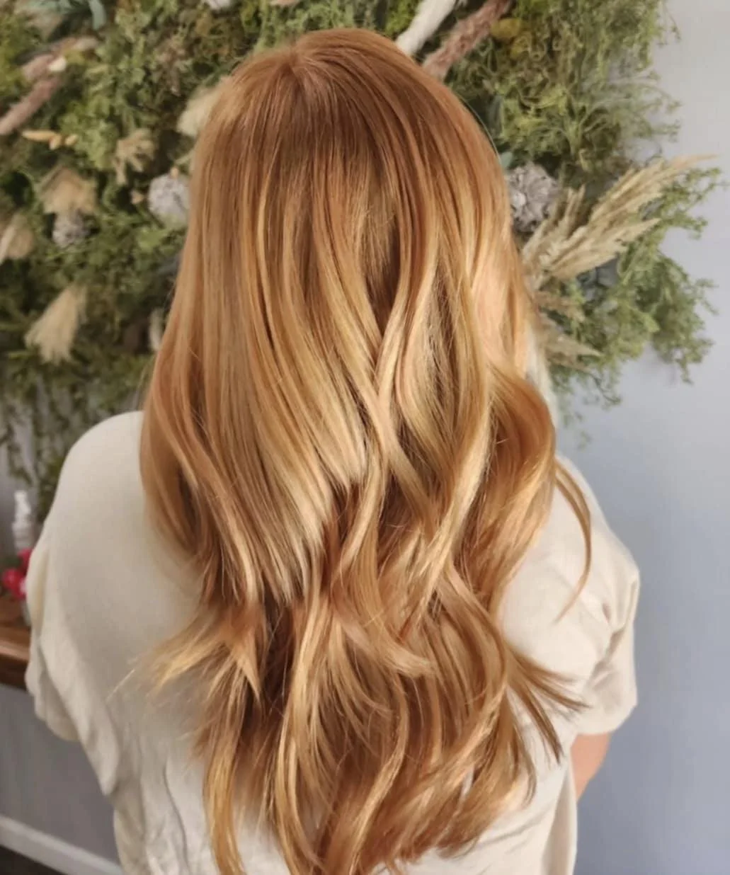 Back view of a woman with long, wavy, blonde hair, standing in front of a floral arrangement.