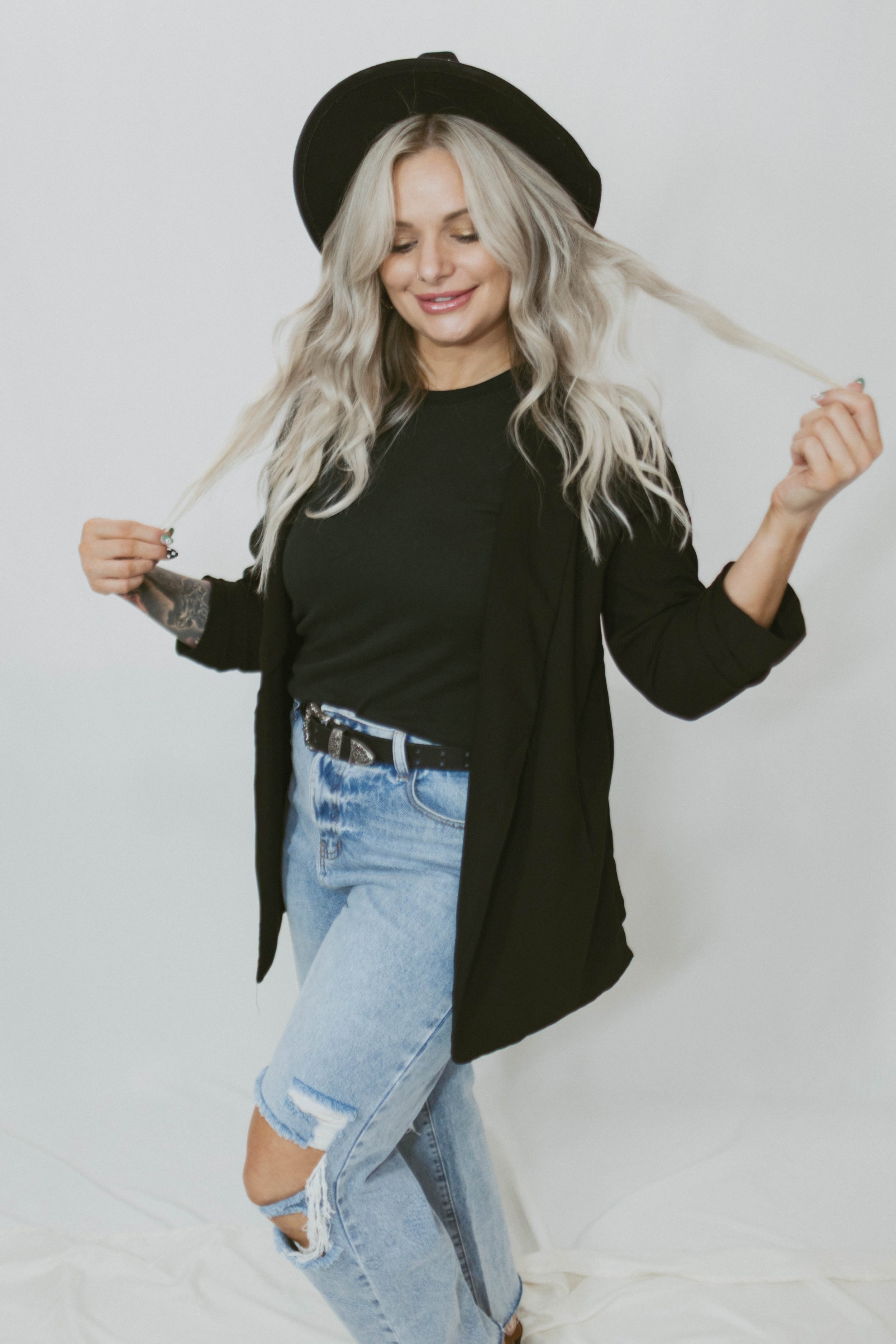 A woman with long blonde wavy hair wearing a black hat, black shirt, black jacket, and ripped light blue jeans. She is smiling and holding her hair with both hands.