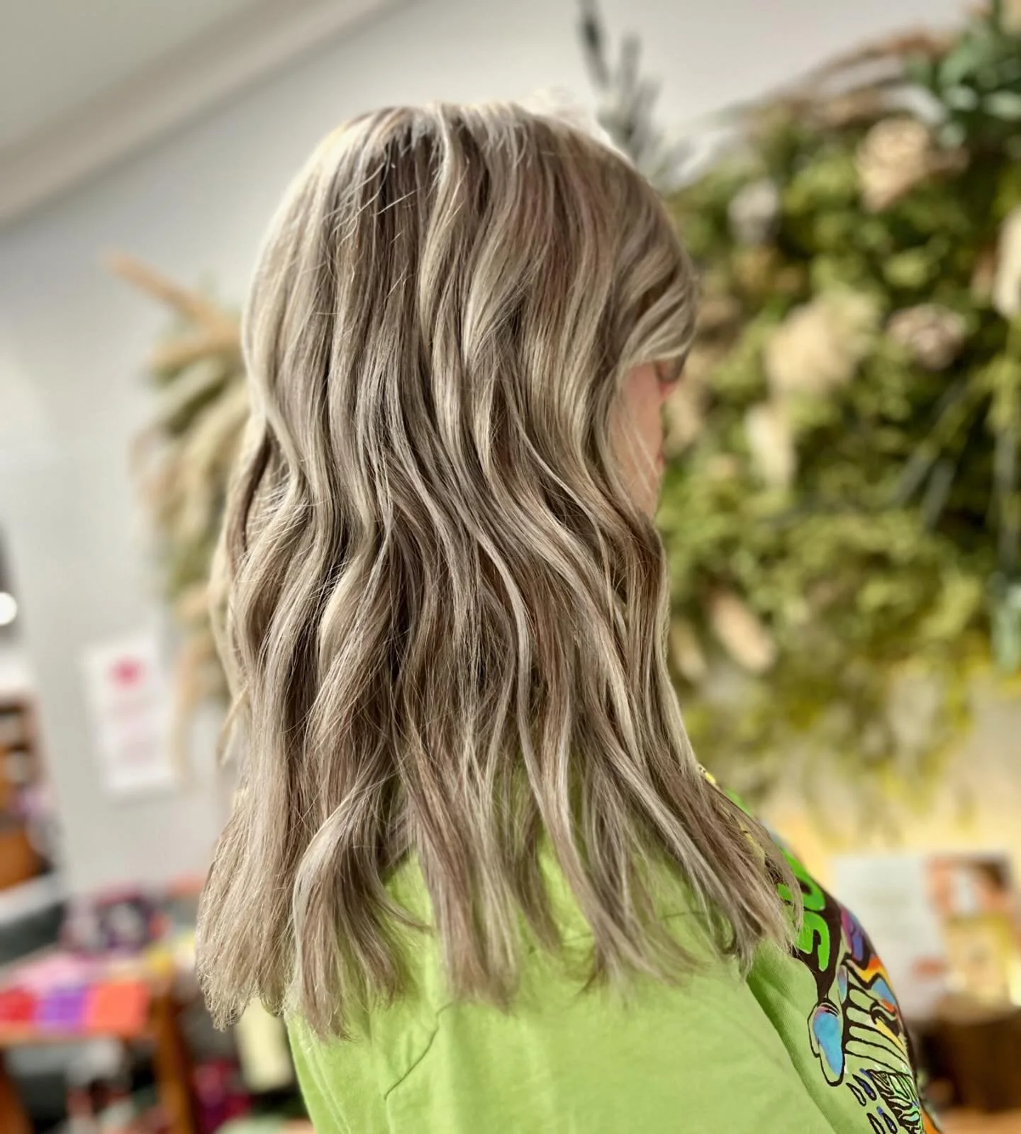 A woman with shoulder-length wavy blonde hair wearing a green shirt, looking away in an indoor setting with plants in the background.