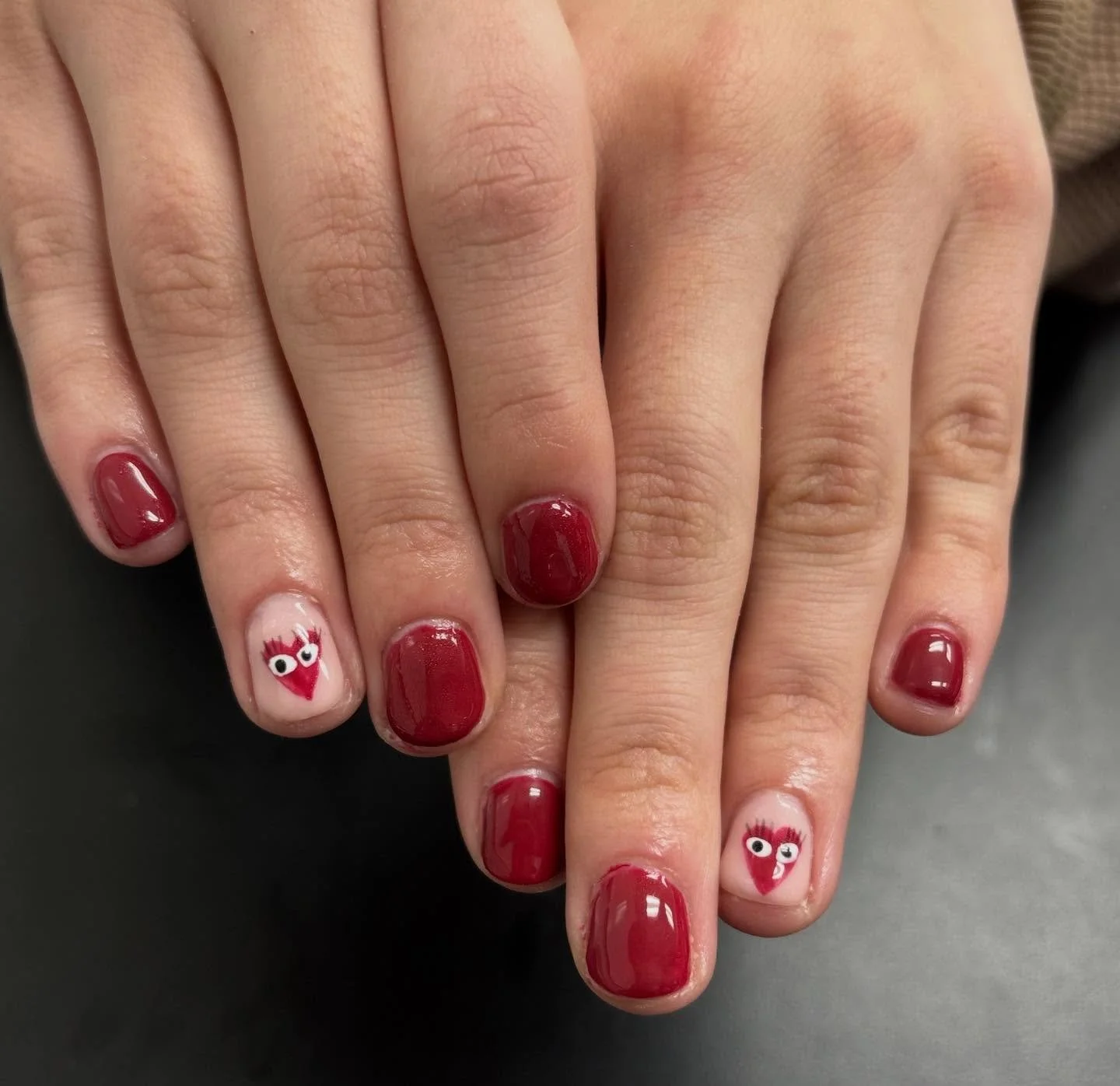 Close-up of a person's hands with red nail polish, featuring cartoonish heart-shaped nail designs with eyes and eyelashes.