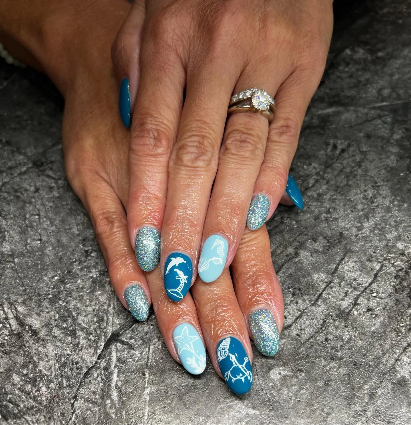 Woman's hands with blue and glittery nail art, an engagement ring with a large gemstone, and a wedding band. The nails feature ocean-themed designs including dolphins, a starfish, and underwater scenery.