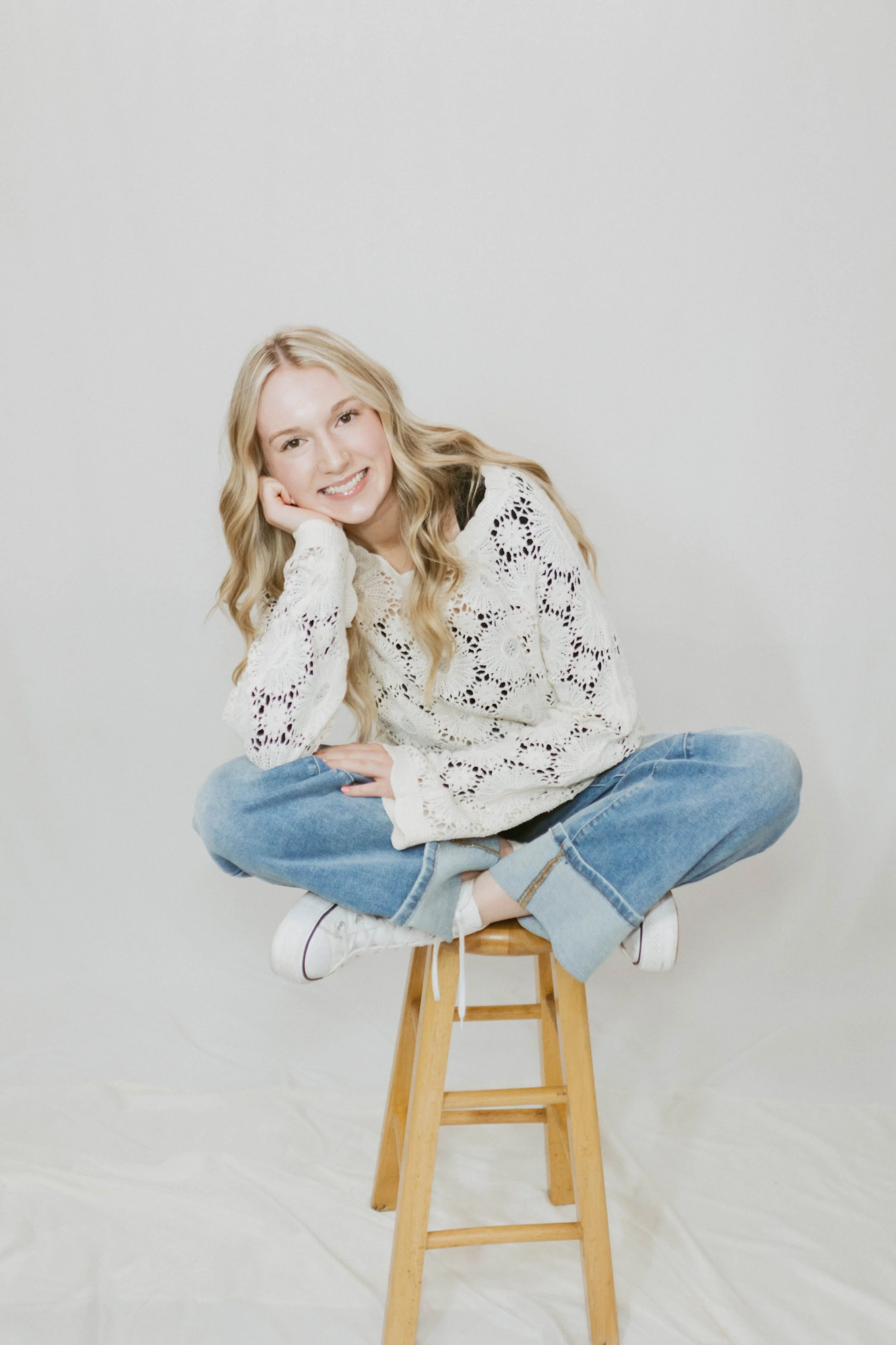 A young woman with long blonde hair sitting cross-legged on a wooden stool, smiling at the camera, wearing a white lace sweater, blue jeans, and white sneakers, against a plain white background.