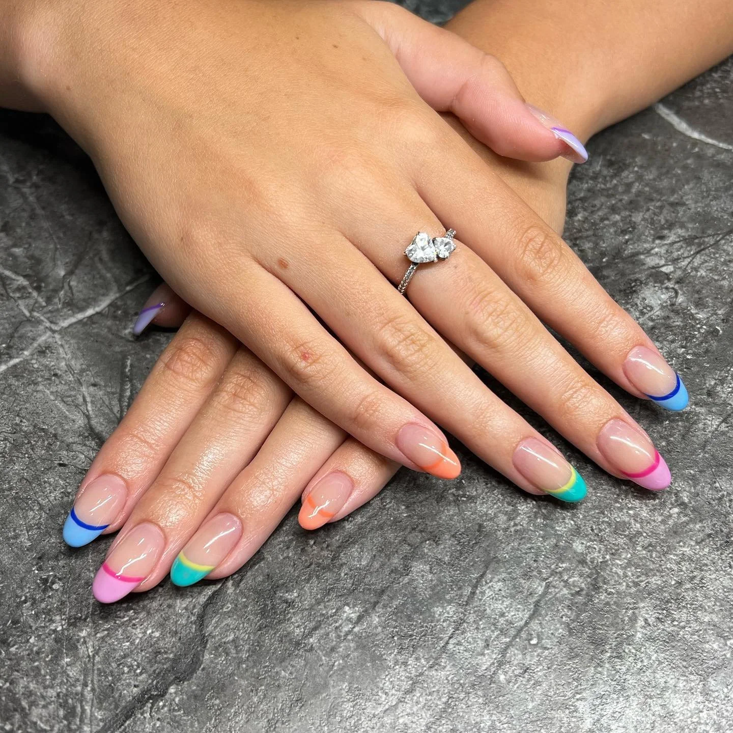 Hands resting on a dark textured surface, one hand displaying a silver ring with a large center stone, surrounded by two smaller stones, and colorful, pastel-painted nails with a French tip design.