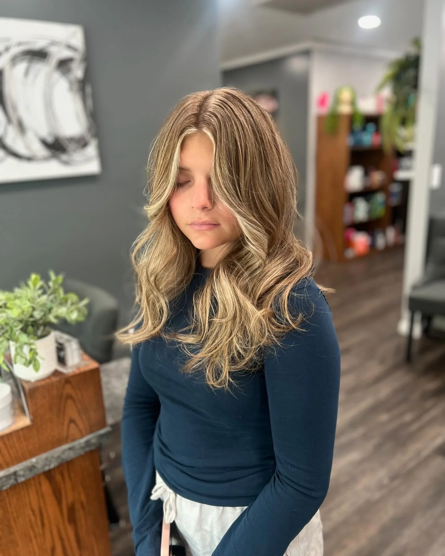 A young woman with long, wavy, blonde hair and light skin, standing indoors with her eyes closed, wearing a navy blue long-sleeved top and light-colored pants, in a living room or salon setting.