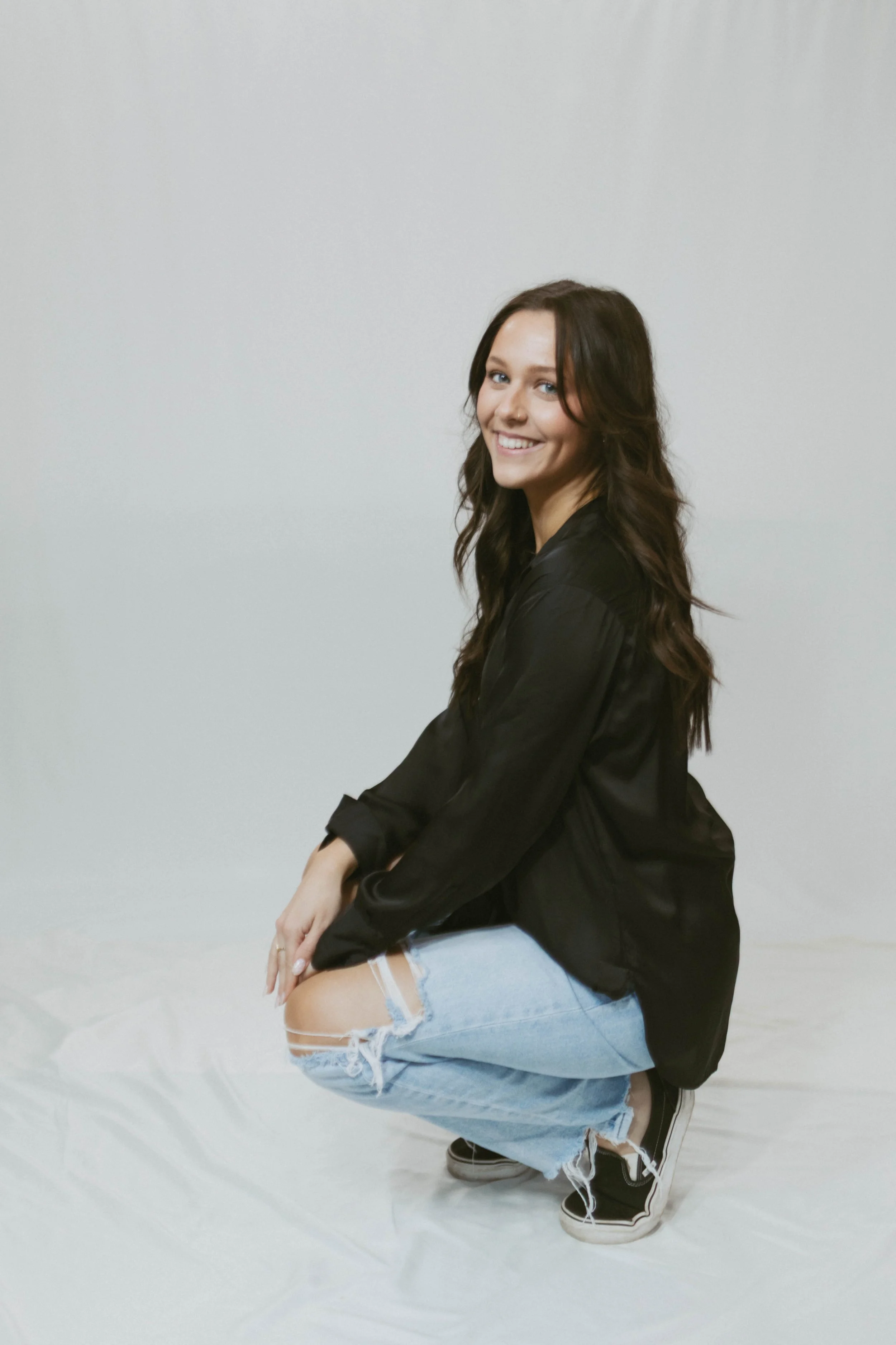 A young woman with long brown hair smiling and squatting in front of a plain light-colored background, wearing a black satin blouse, ripped jeans, and black sneakers.