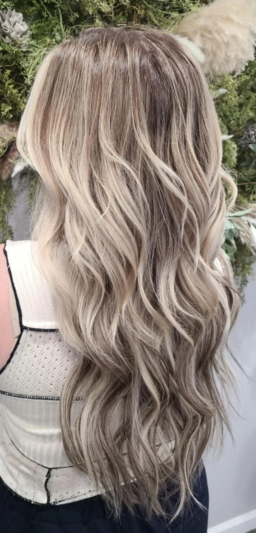 Back view of a woman with long, wavy blonde hair, with highlights, standing outdoors near greenery and white flowers.