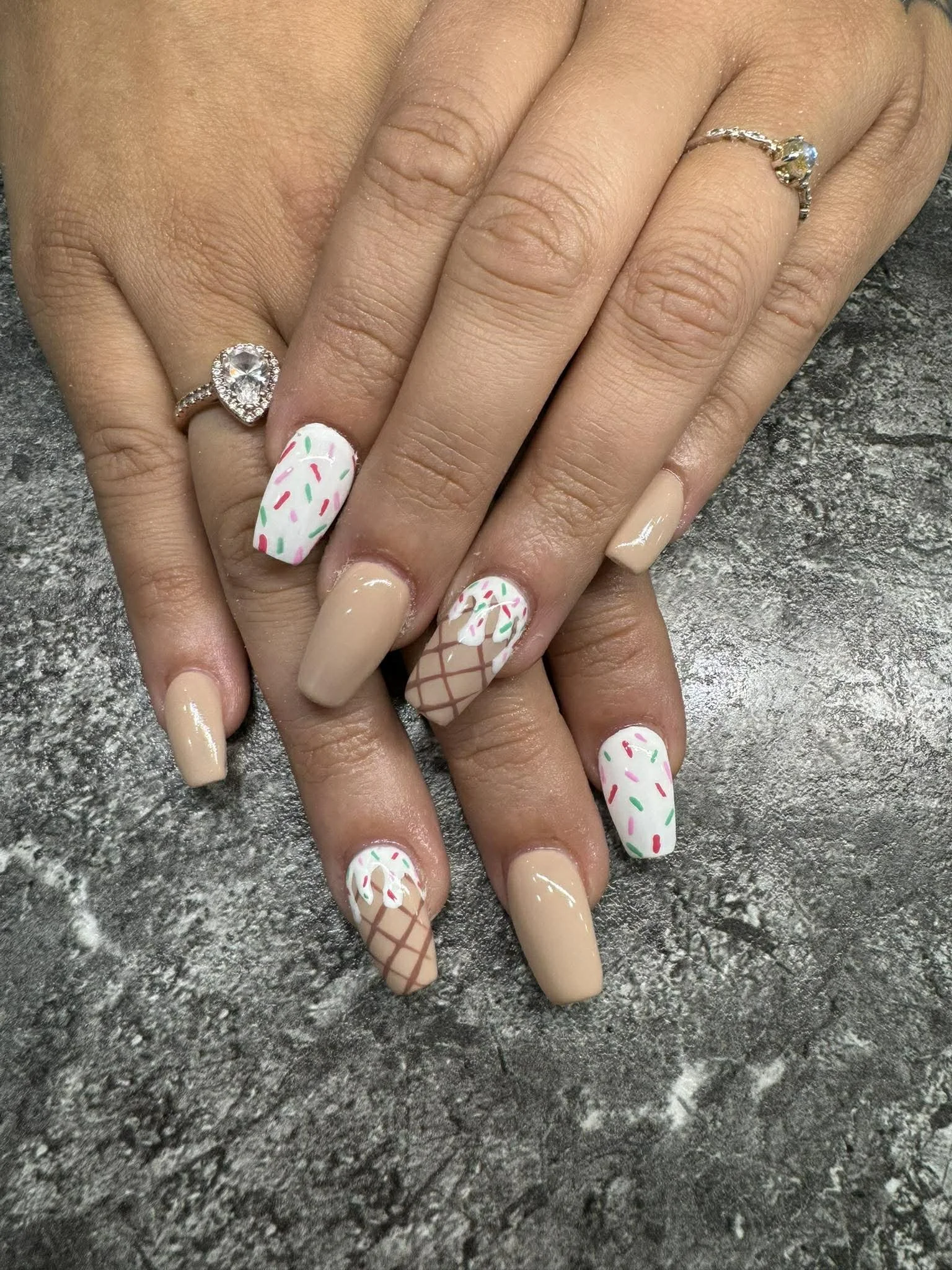 Hands with beige and decorated nail polish, one hand wearing two rings, one with a large clear stone, and the other with multiple smaller stones.