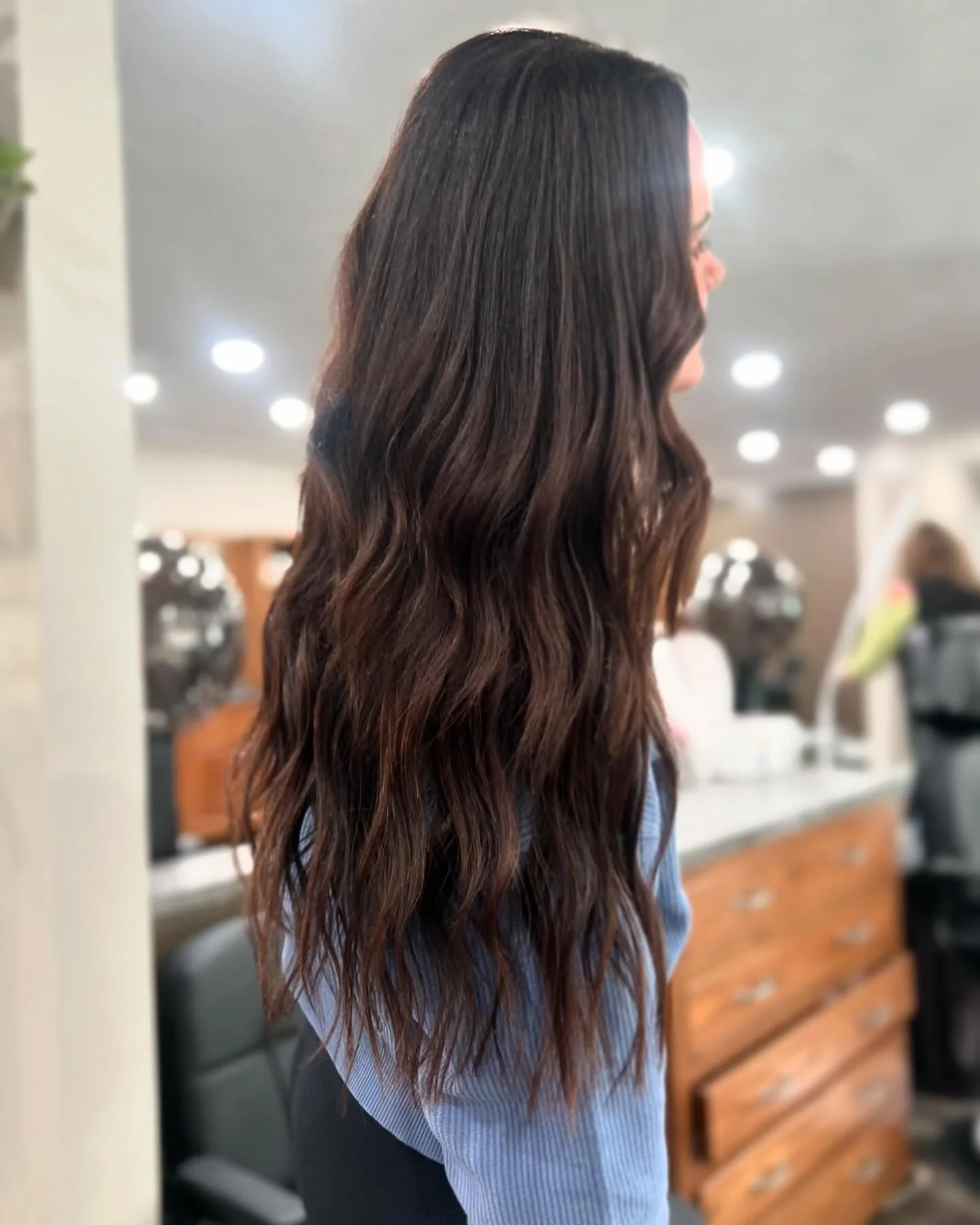 Side view of a woman with long, wavy brown hair in a salon or indoor setting.