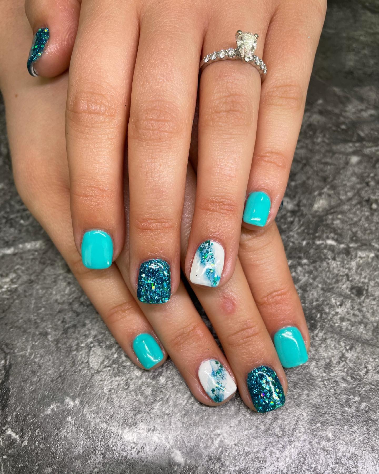 Manicured hands with teal, white, and glittery blue nail polish, one ring with a large stone on ring finger.