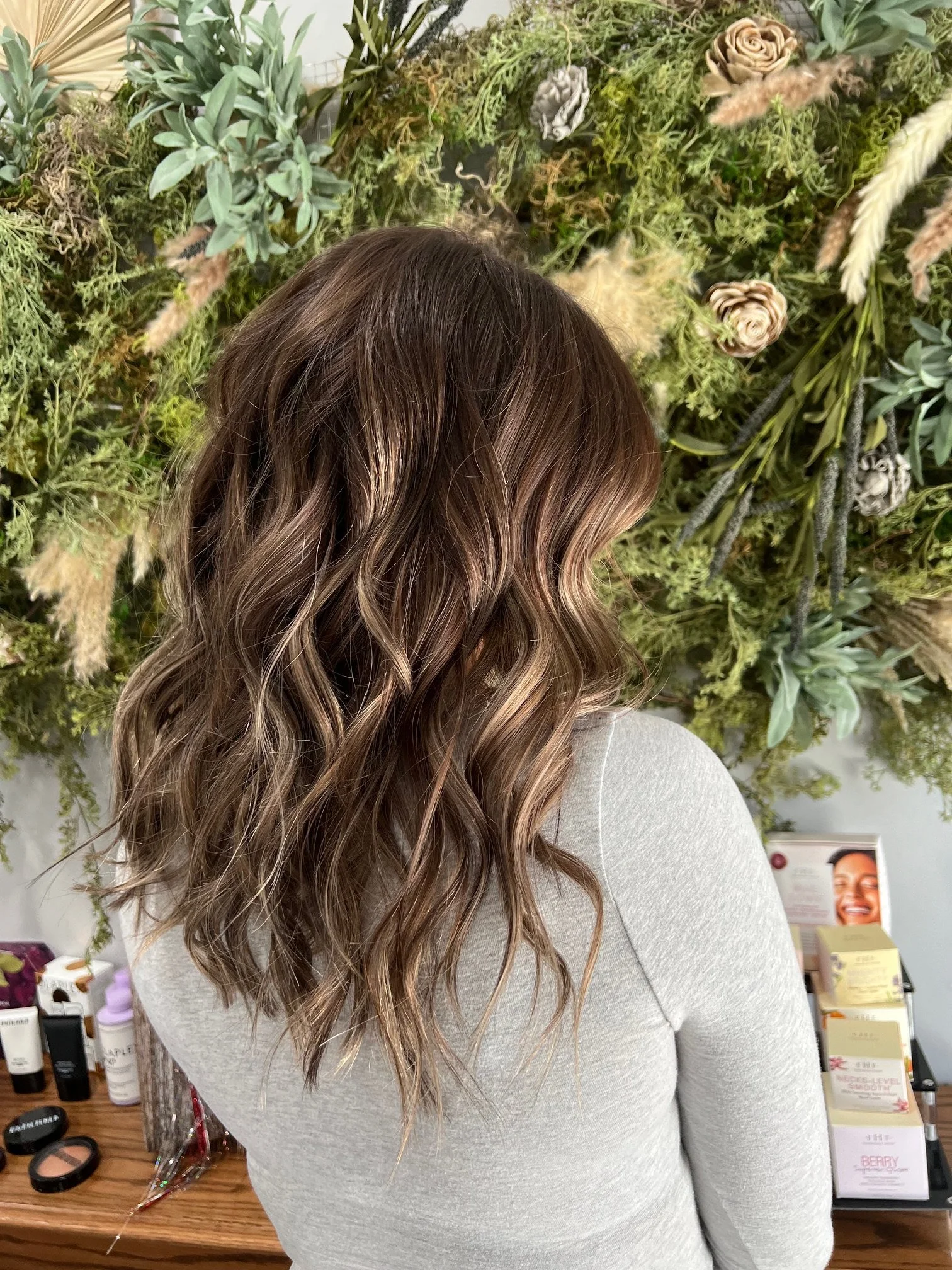 A woman with wavy brown hair standing in front of a greenery wall with various plants and flowers.