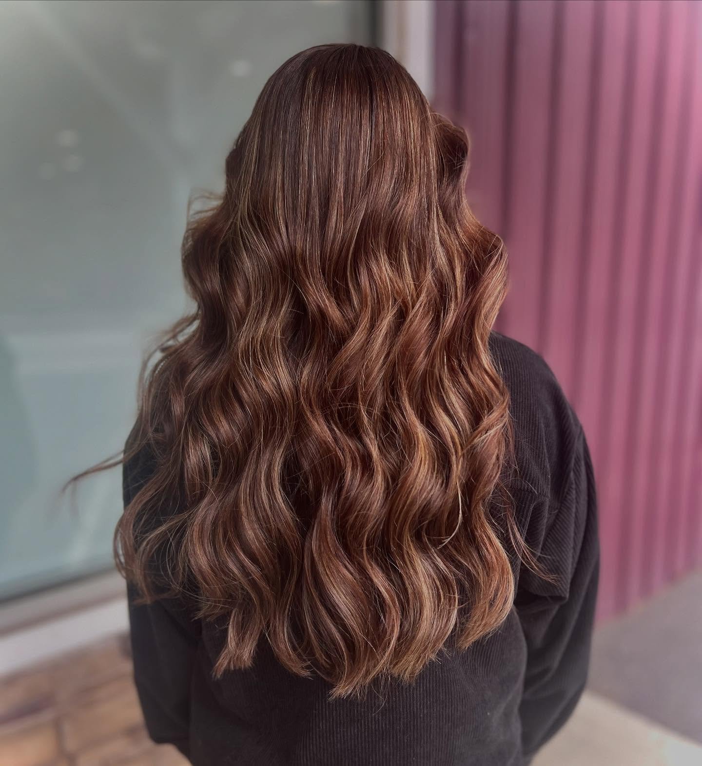 Back view of a woman with styled, wavy, chestnut brown hair, wearing a black top, standing in front of a purple striped wall.