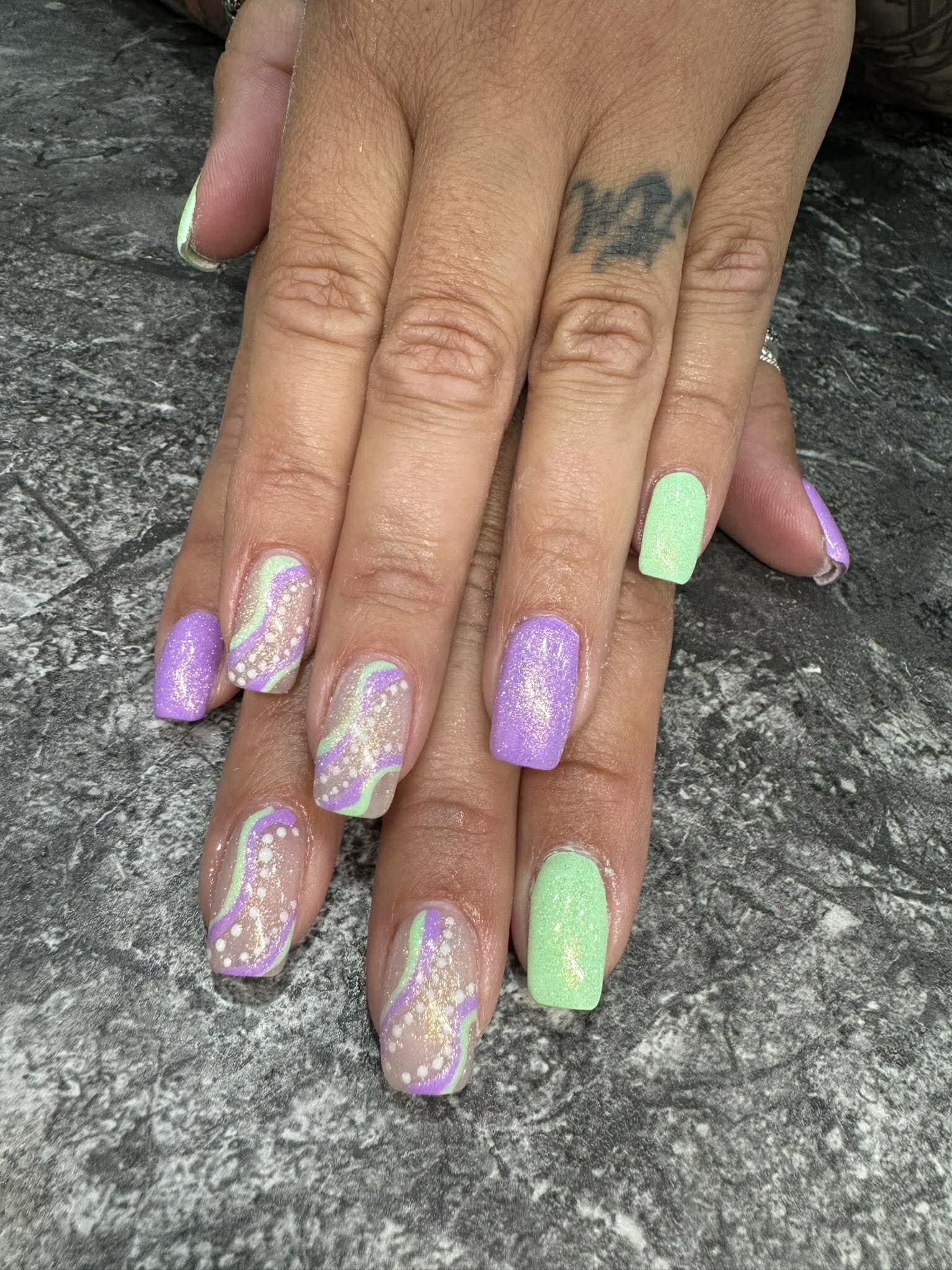Woman's hands with pastel-colored, glittery, marbled nail polish on her nails, resting on a textured dark gray surface. She has a tattoo on her finger.