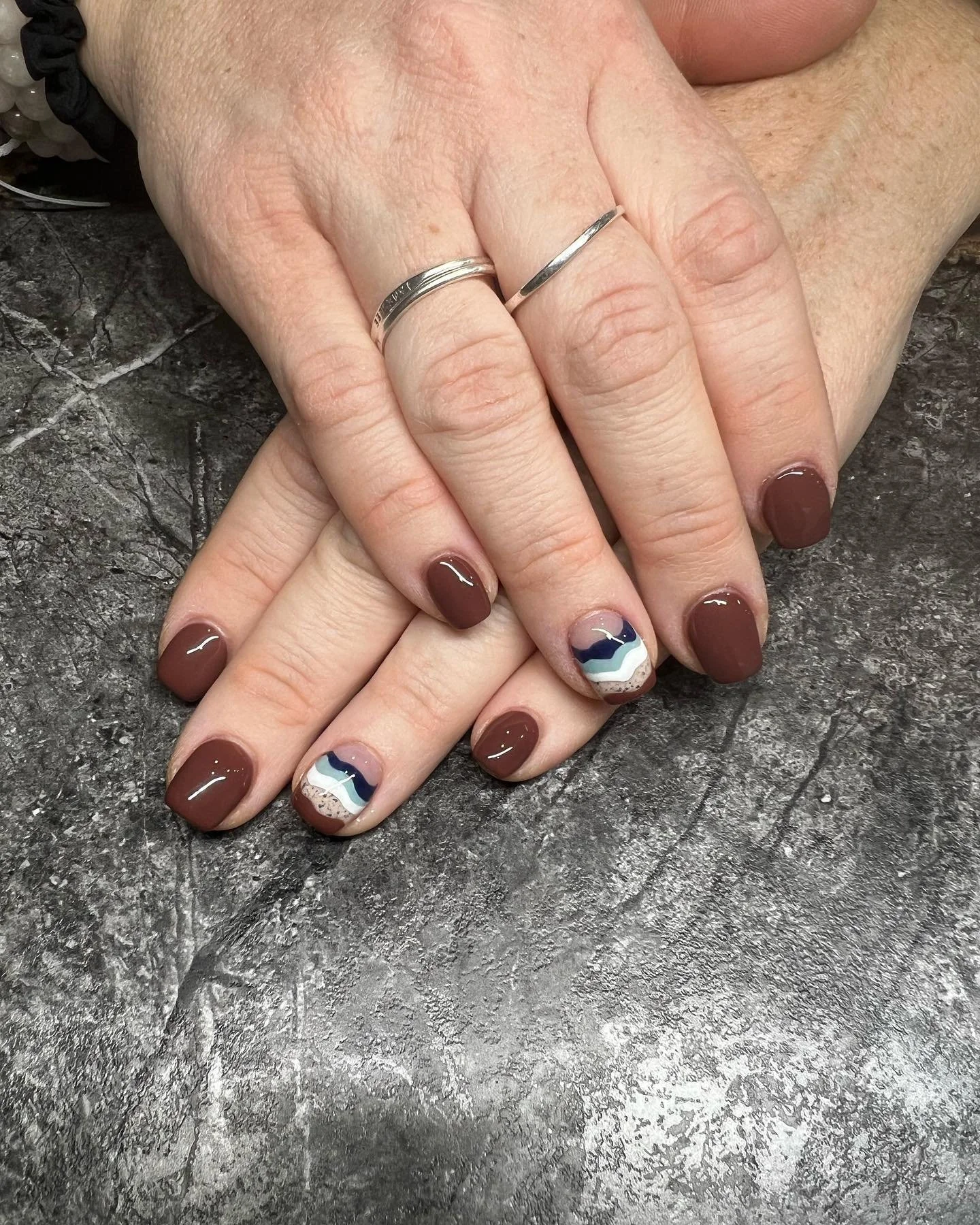 Close-up of two hands with manicured nails, one with brown polish and the other with a colorful, wavy design. The hands are resting on a stone surface, and there are two silver rings on the fingers.