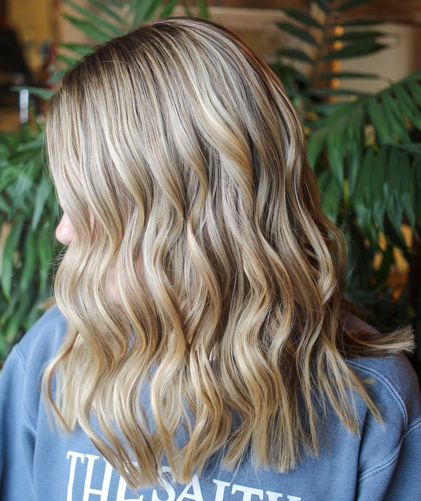 Close-up of wavy blonde hair on a person wearing a blue shirt with text, with green plants in the background.