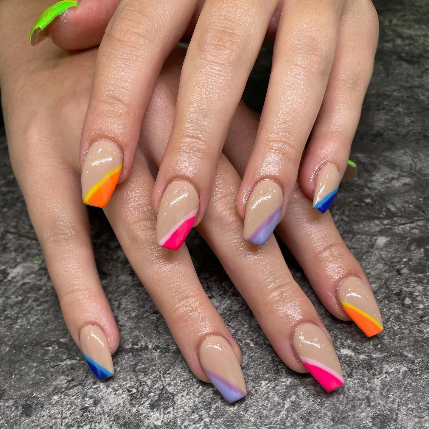 Nude-colored manicured nails with colorful diagonal tips in shades of pink, orange, yellow, purple, and blue, resting on a dark textured surface.