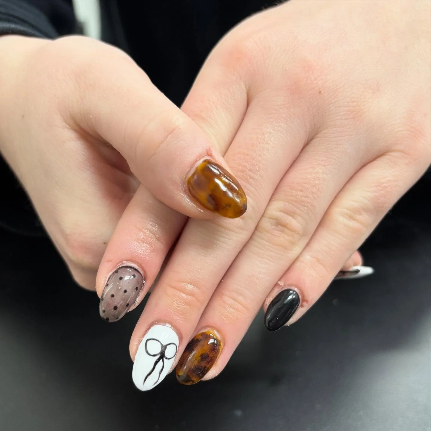 Close-up of a hand with unique manicured nails featuring various designs and colors, including black, white with a bow, beige with black dots, and tortoiseshell pattern.