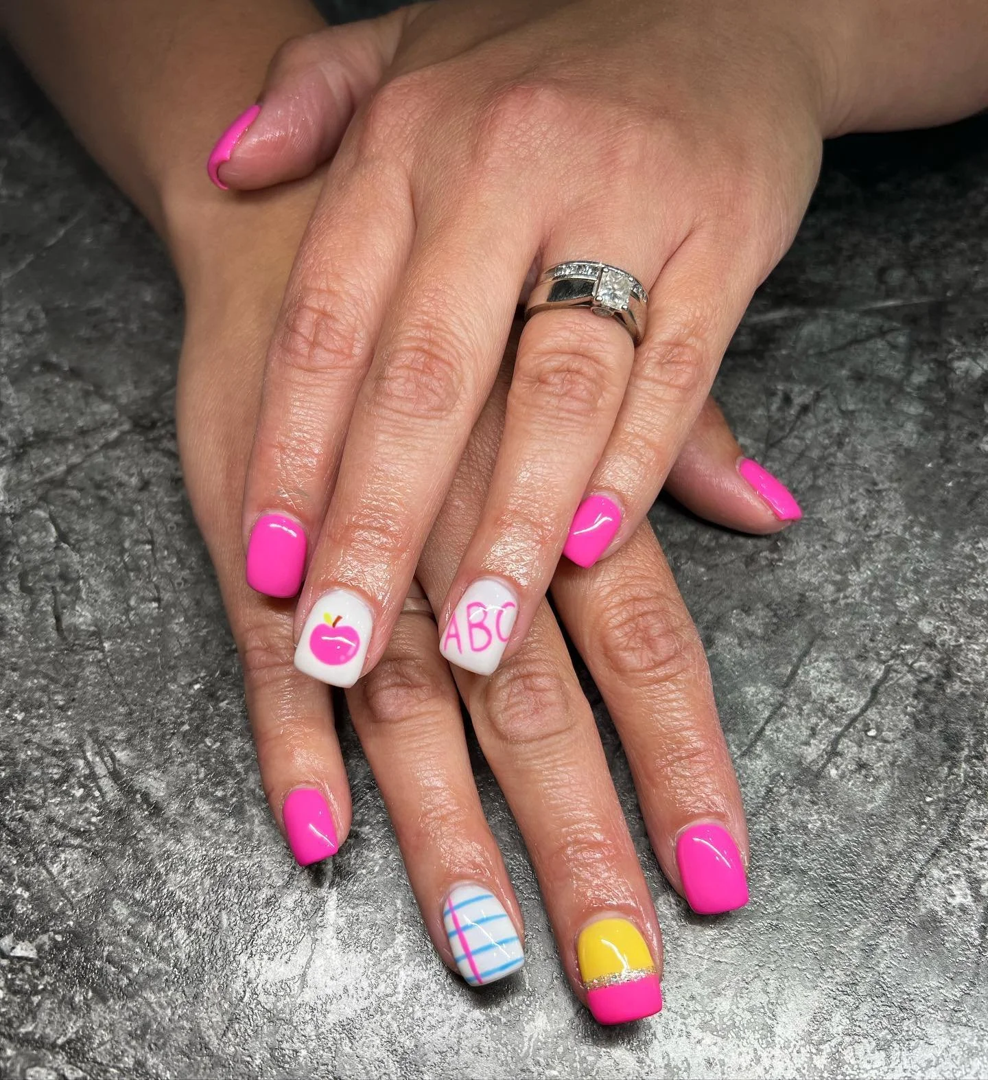 Nail art on hands with pink, yellow, and blue nails, featuring apple, ABC, and striped designs, jewelry including rings, and a gray textured surface.