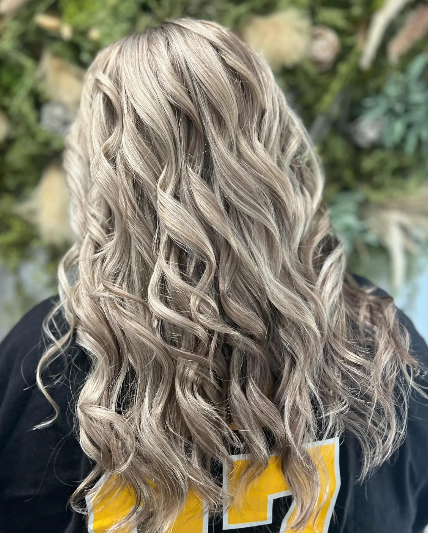 Back of head with long, blonde, curly hair, outdoors, with blurred greenery background.