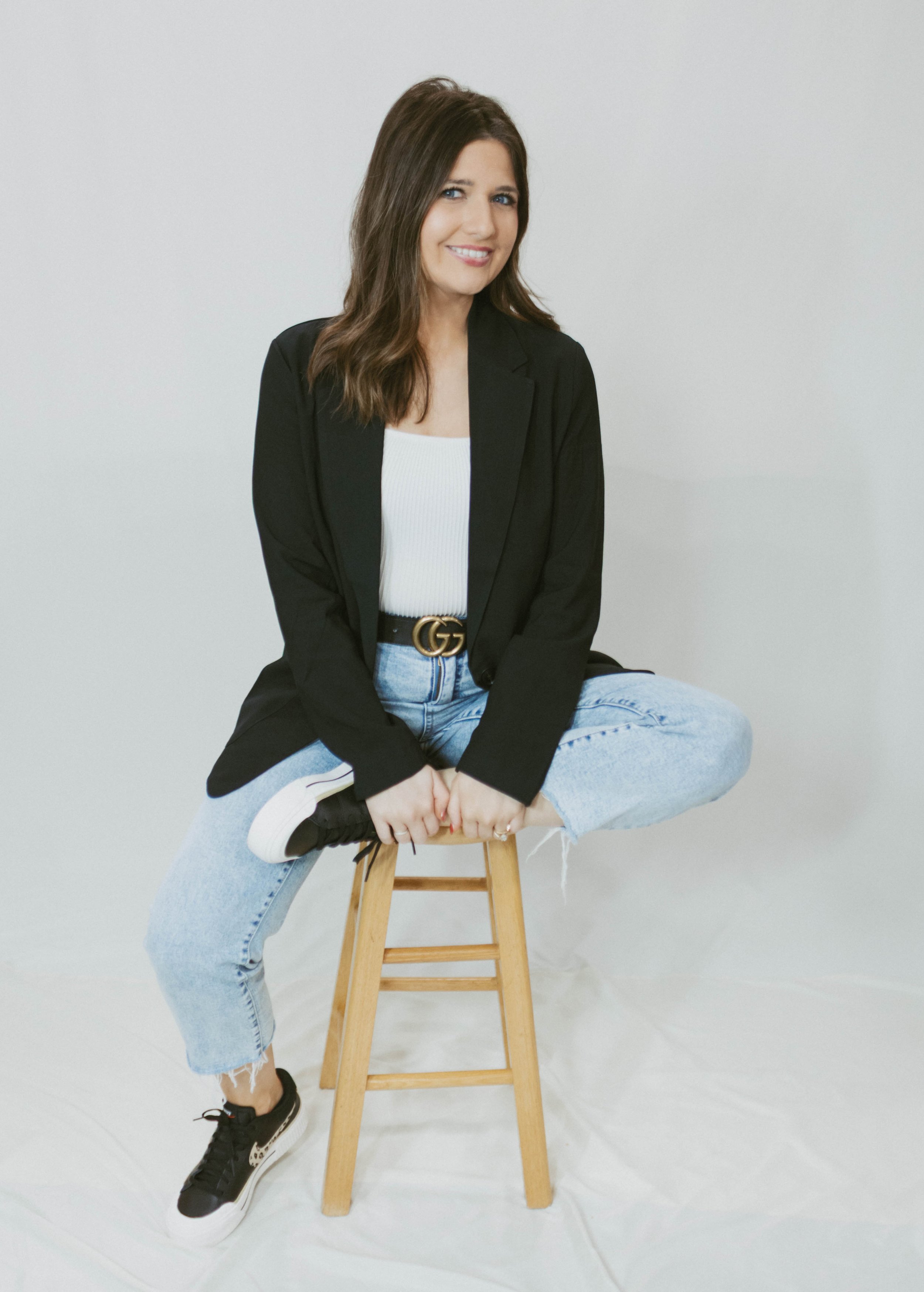 A woman with brown hair sitting cross-legged on a wooden stool, wearing a black blazer, white top, light blue jeans, and black sneakers, against a plain white background.