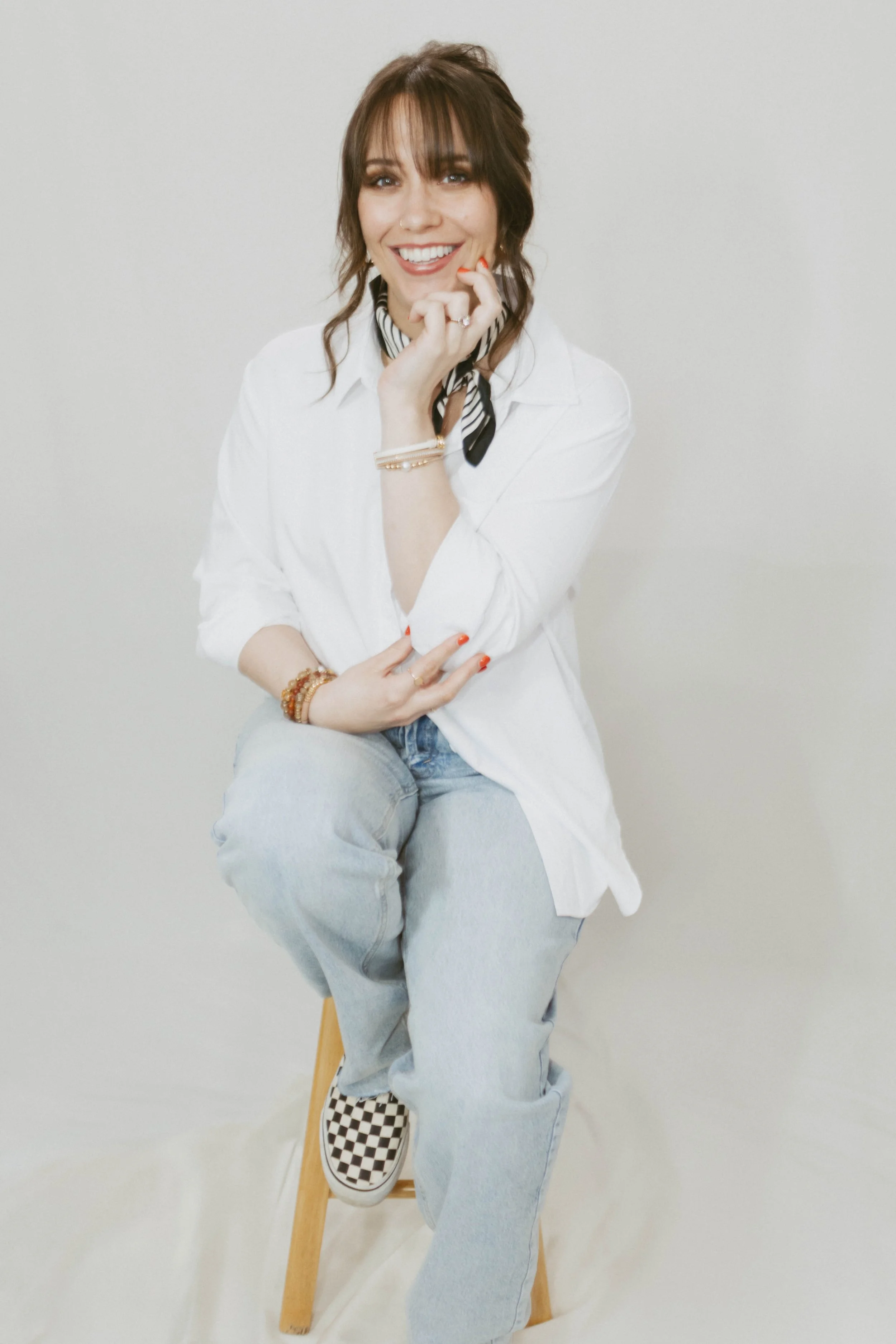 A woman with brown hair, smiling, wearing a white shirt, a black and white striped scarf, light blue jeans, and checkerboard shoes, sitting on a wooden stool against a plain light-colored background.