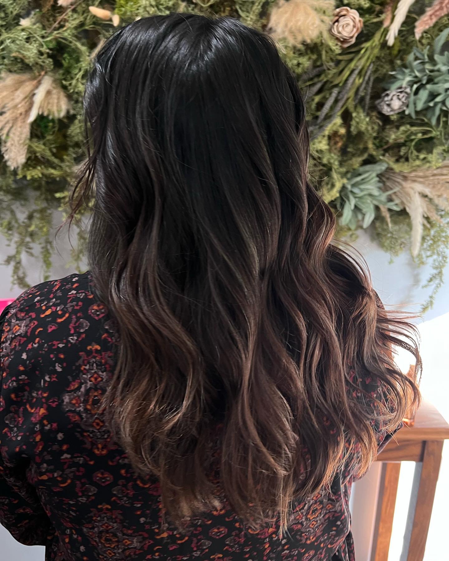 Back view of a woman with long wavy dark hair with brown highlights, wearing a dark floral blouse, standing in front of a wall decorated with dried flowers and greenery.