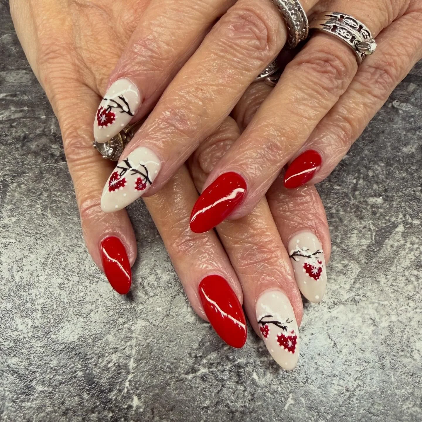 Hand with red and white Christmas-themed nail art, featuring hearts, snowflakes, and tree designs, against a gray textured surface.