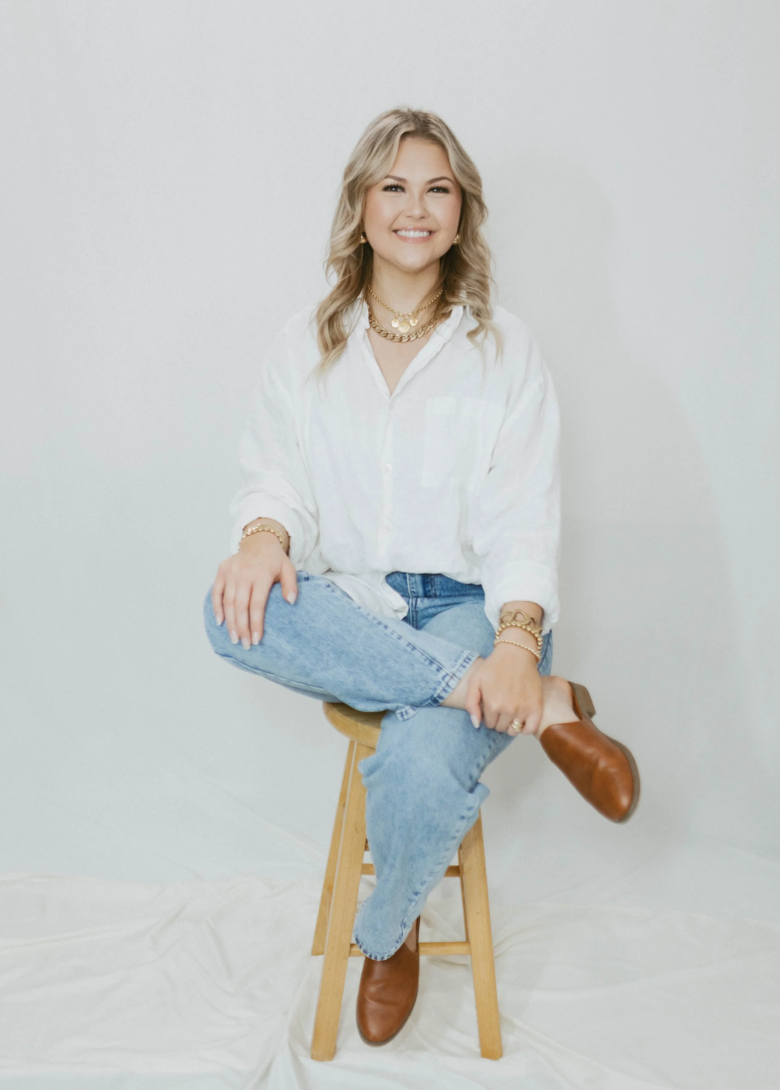 A young woman sitting on a wooden stool against a plain white background, smiling at the camera. She has blonde, wavy hair and is dressed in a white button-up shirt, light blue jeans, and brown ankle boots. She is accessorized with layered gold necklaces, bracelets, and earrings.