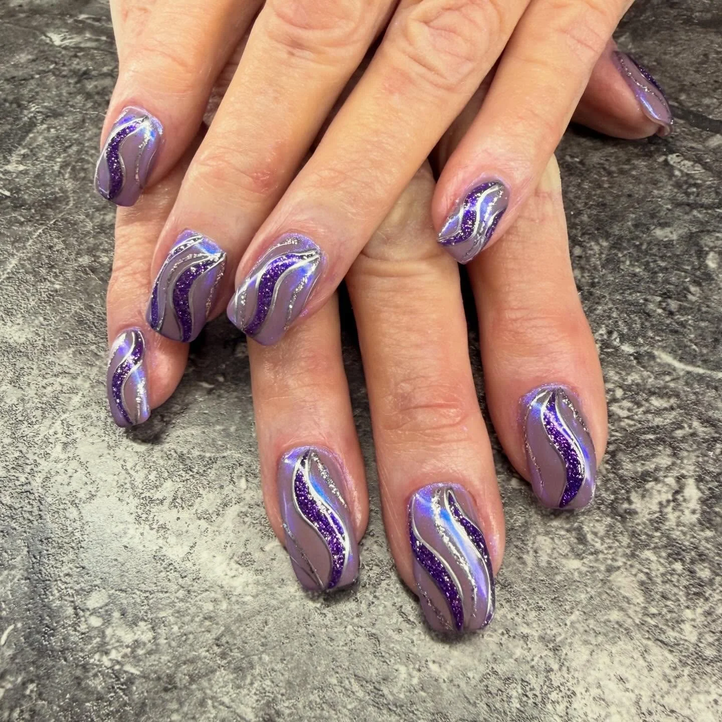 Close-up of hands with purple, silver, and blue glitter swirl nail polish on manicured nails, resting on a textured gray surface.