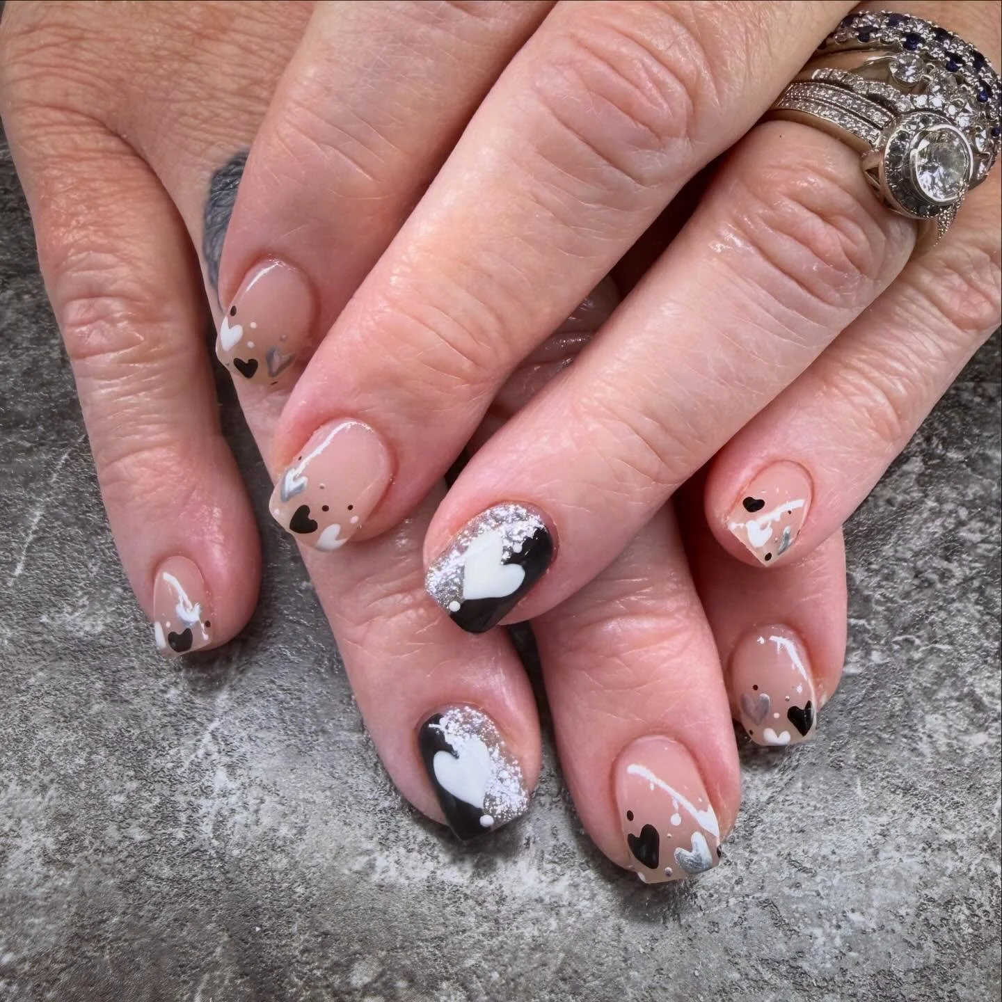 Close-up of hands with decorated nails featuring a black and white heart design, glitter accents, and a neutral base color. The person is wearing multiple rings, including a large diamond ring.