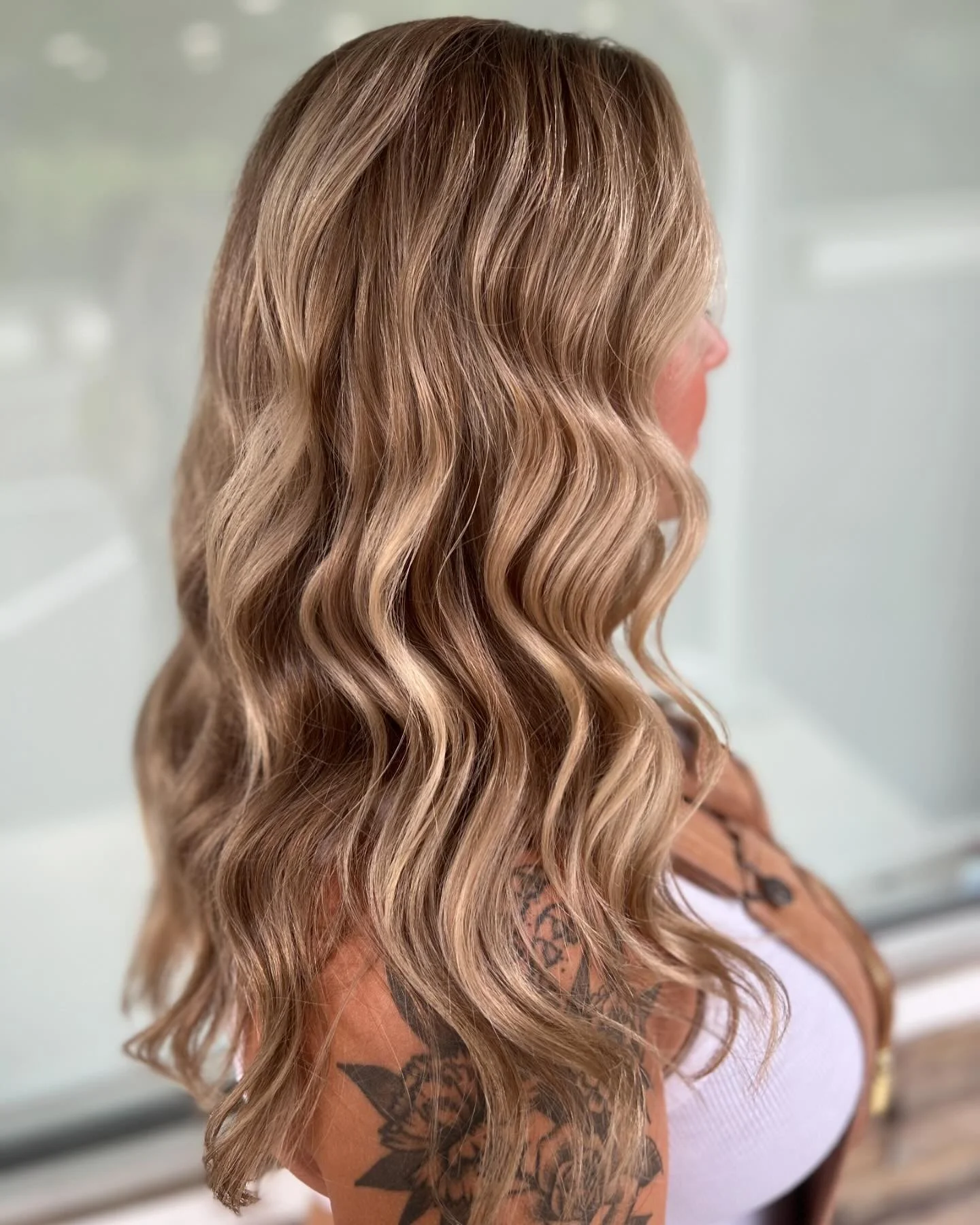 Side view of a woman with long, wavy, blonde hair, wearing a white top and a brown vest, showing tattoos on her shoulder and arm.