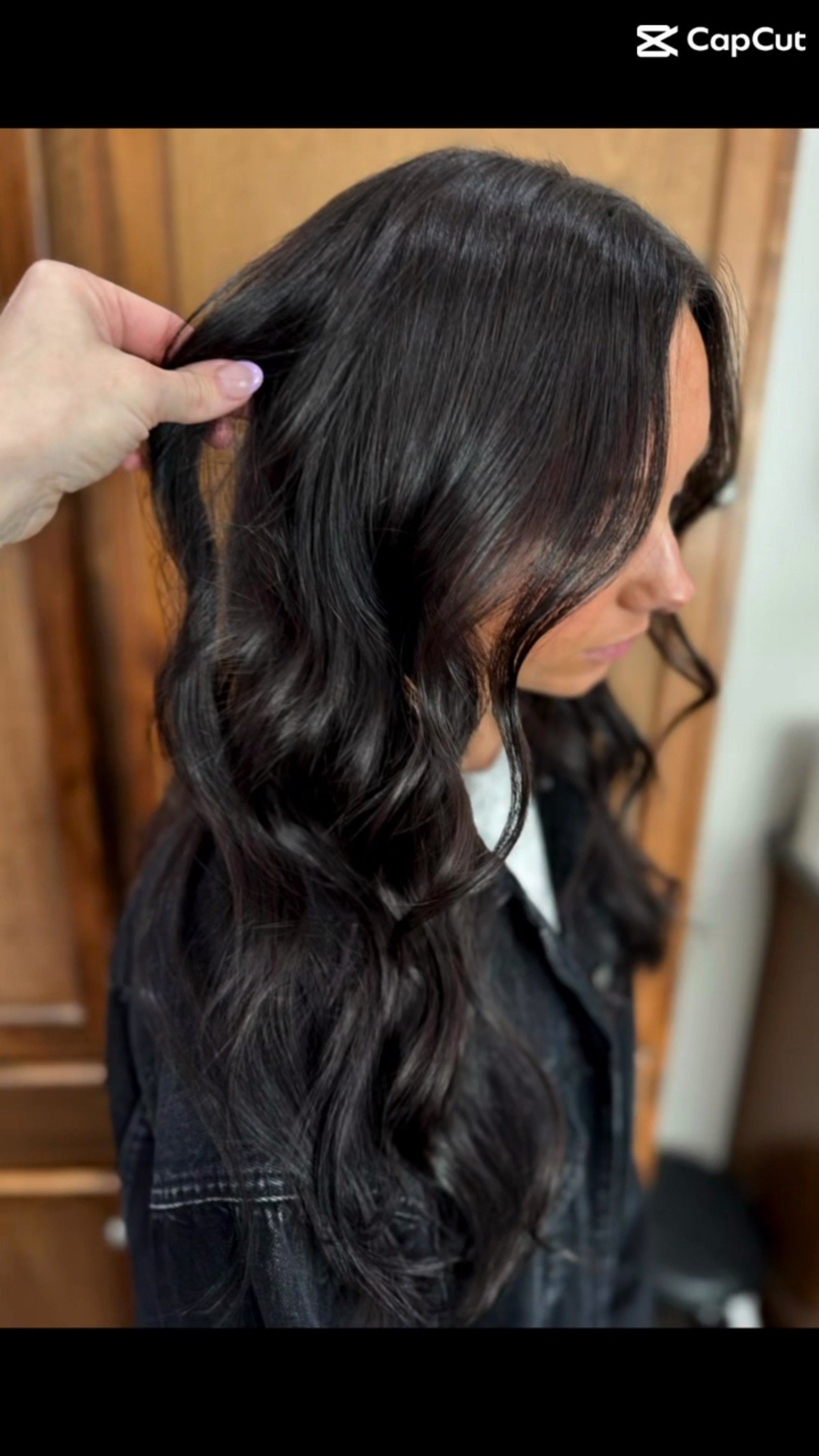 A woman with long, dark, wavy hair being styled or curled, with someone holding a section of her hair.