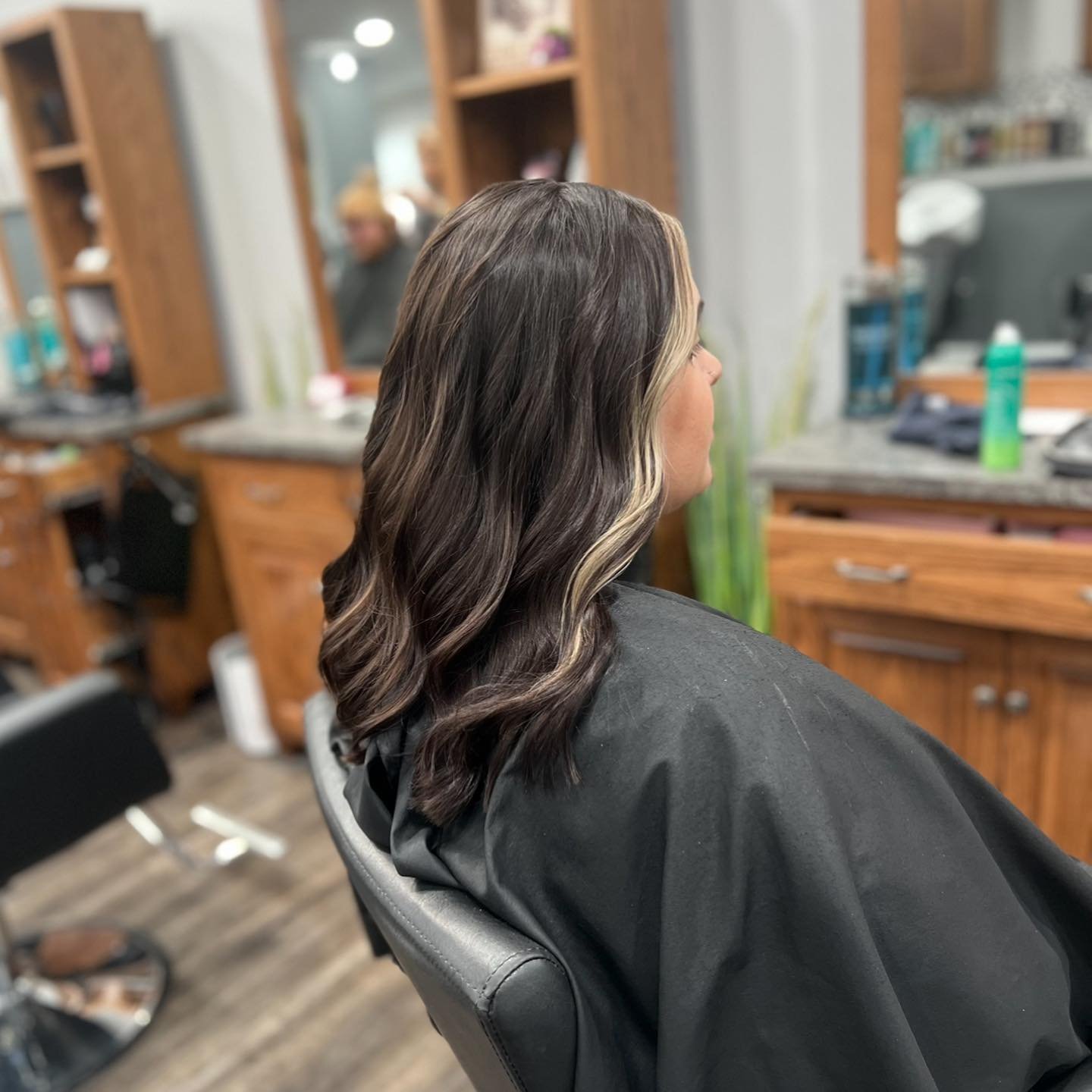 A woman with long dark hair with blonde highlights sits in a salon chair, waiting to get her hair styled. The salon interior includes wooden cabinets, a mirror, and various hair products and tools visible on the countertop.