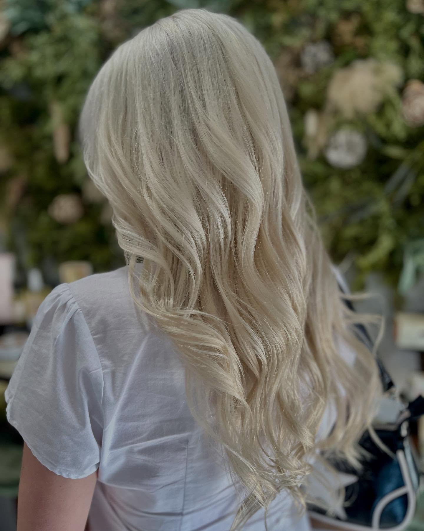 A woman with long, wavy, platinum blonde hair wearing a white shirt, standing indoors in front of a blurred green foliage background.
