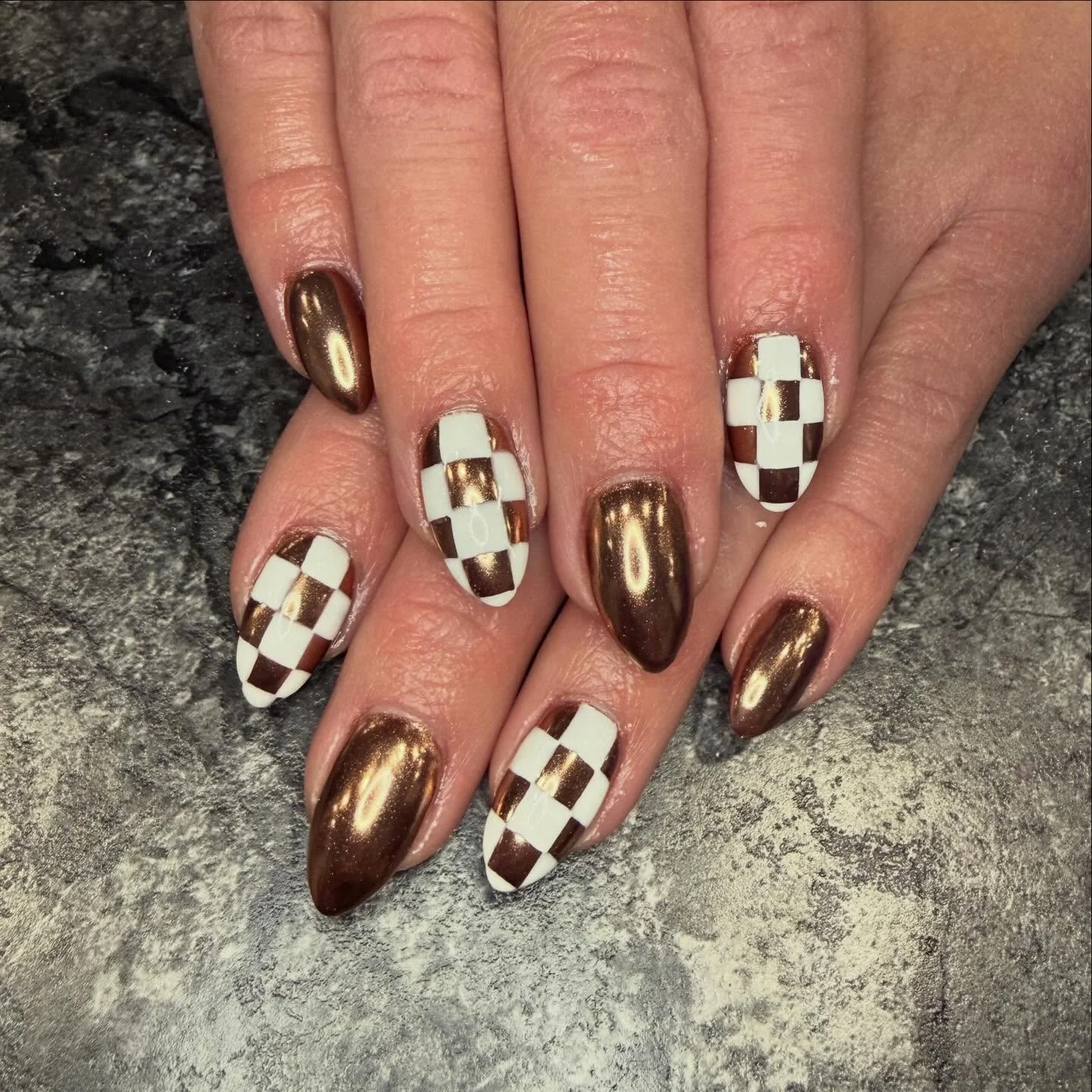 Manicured nails with a checkered pattern in white and brown, some nails painted with metallic bronze polish, on a dark textured surface.